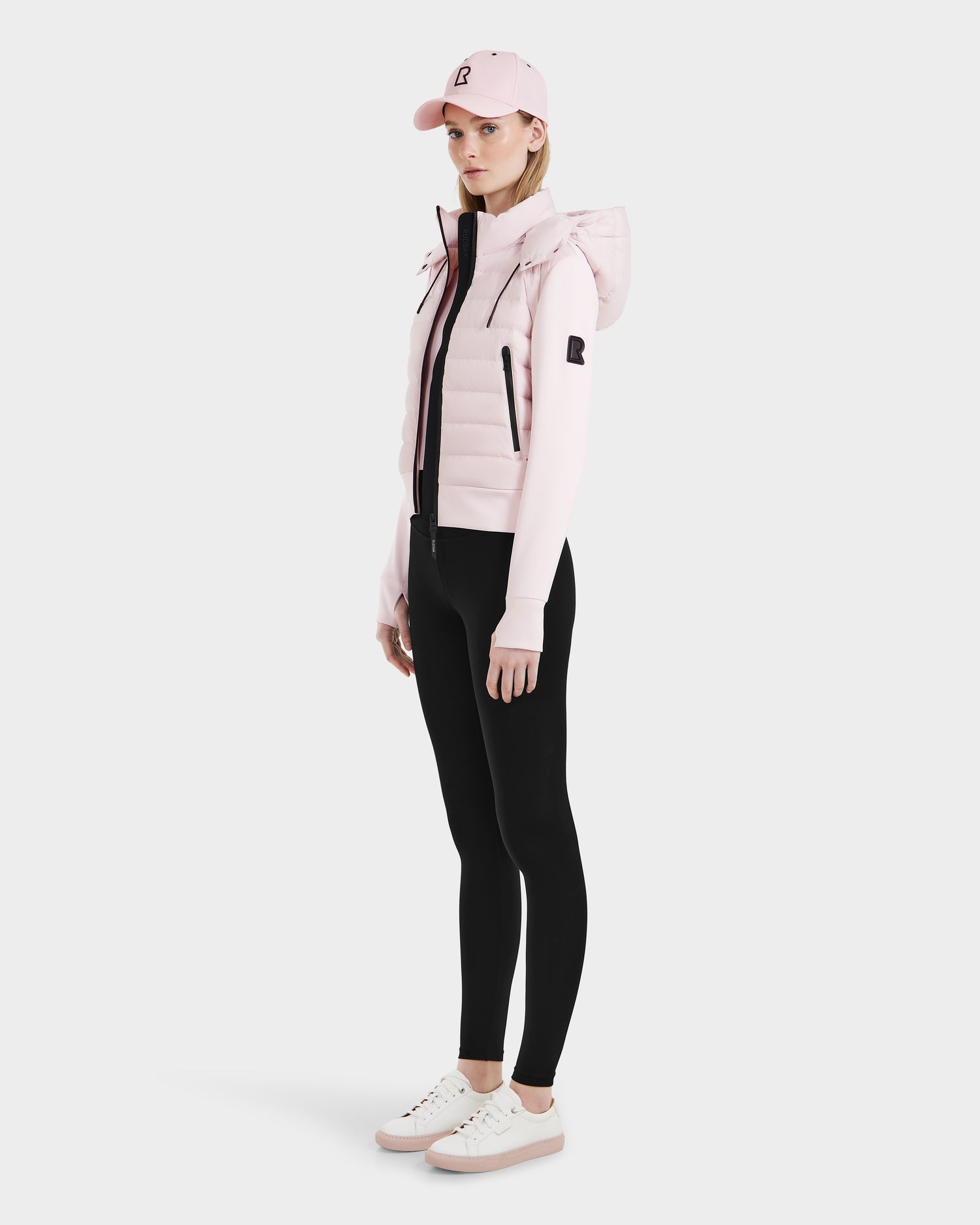 A woman stands sideways against a plain backdrop, wearing the Rudsak CASSA - 8125057 light pink cropped jacket with a matching pink cap, black leggings, and white sneakers.