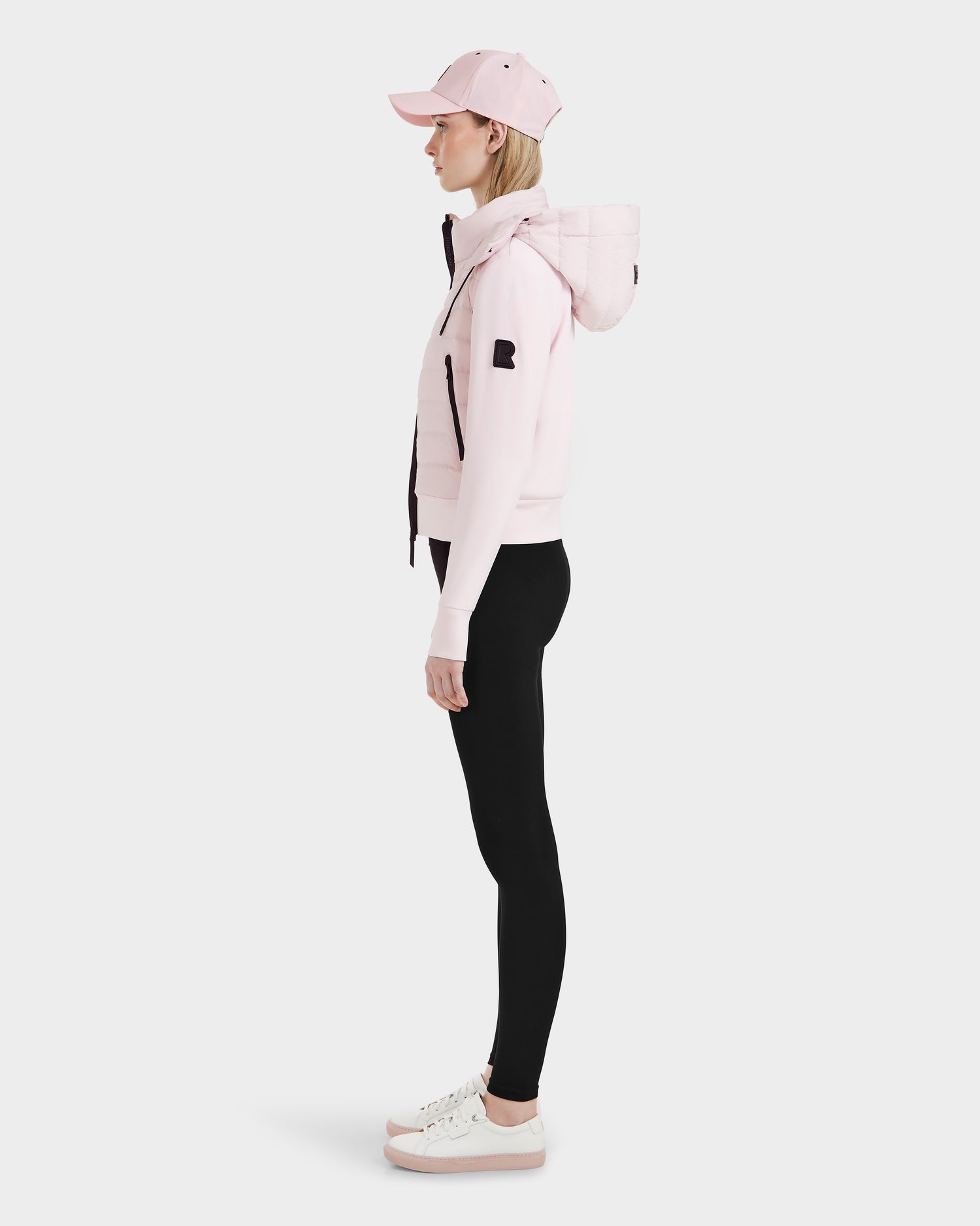 A woman stands in profile against a plain white background wearing the Rudsak CASSA - 8125057 lightweight cropped jacket in light pink, black leggings, pink and white sneakers, and a pink baseball cap.