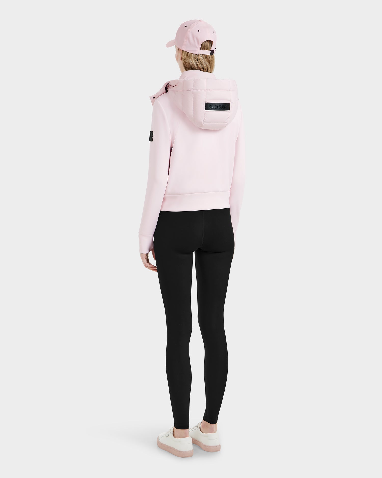 A person stands facing away, wearing the Rudsak CASSA - 8125057 hoodie in light pink, paired with a pink cap, black leggings, and pink sneakers against a plain white background.