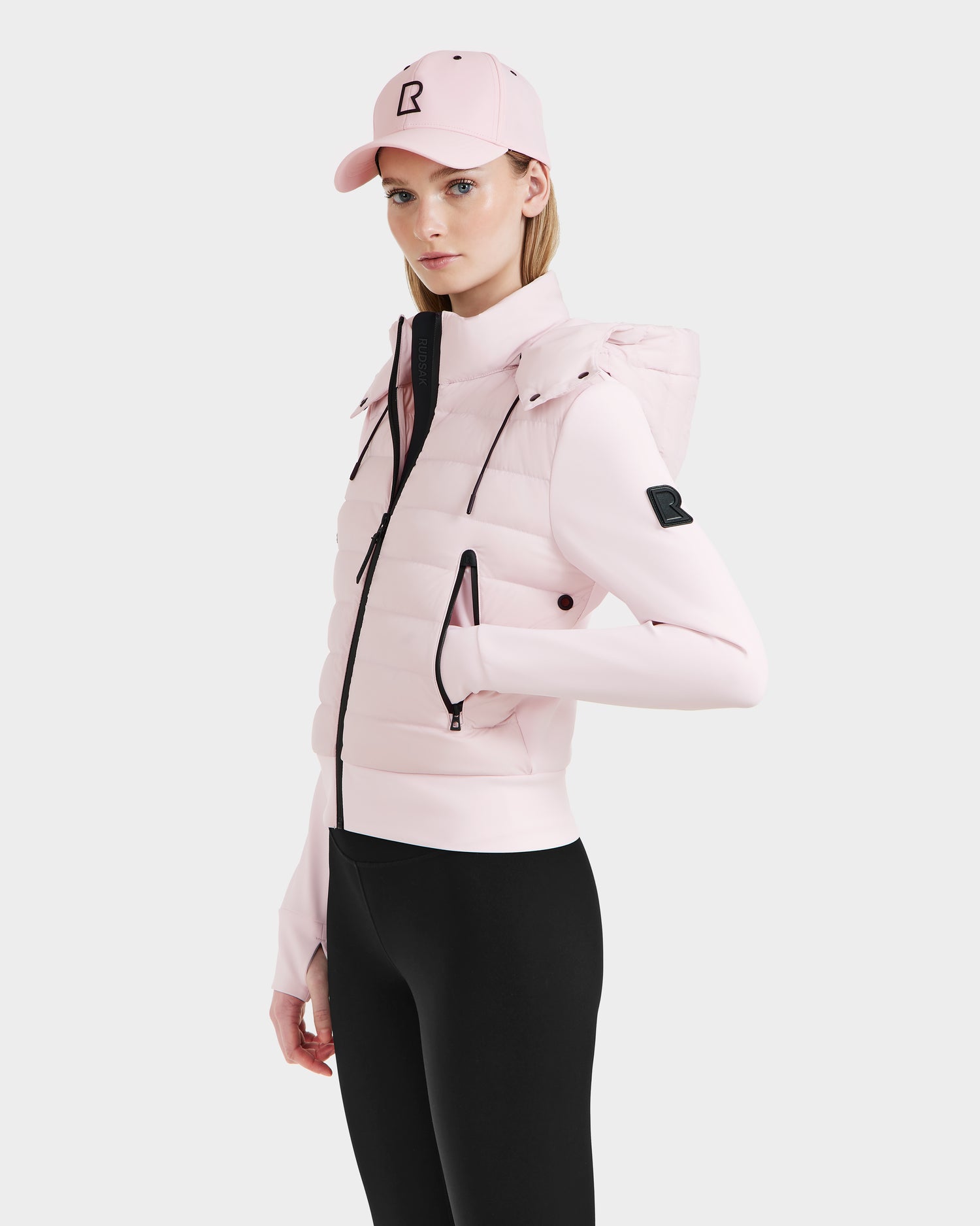 A woman in the Rudsak CASSA - 8125057 Light Pink transitional jacket with a hood, paired with a matching light pink baseball cap and black leggings, stands against a plain white background.