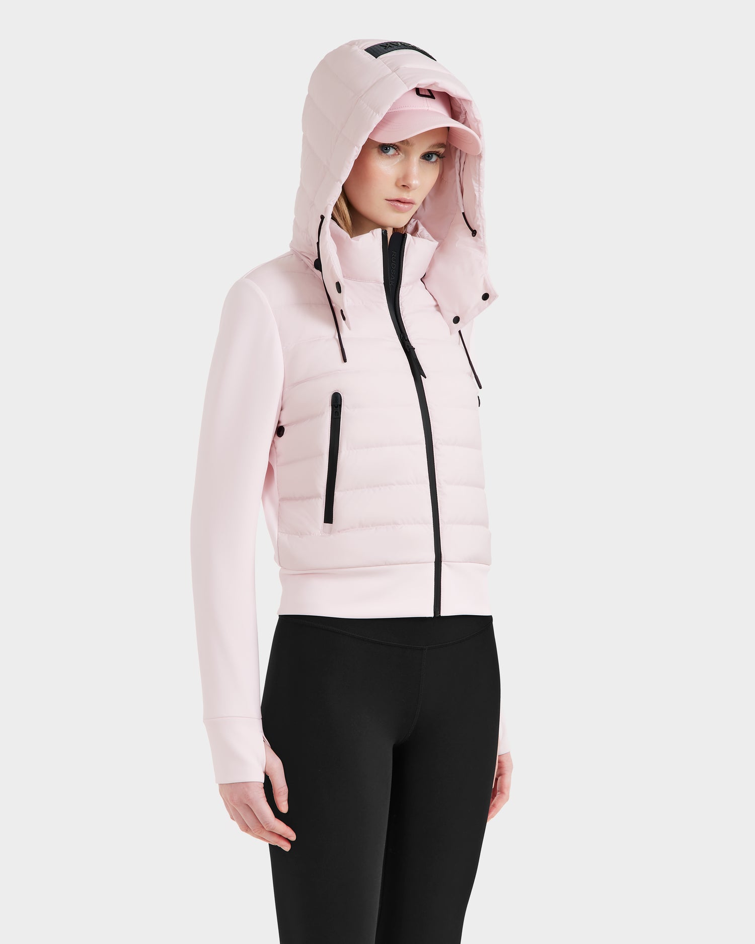 A woman models the Rudsak CASSA - 8125057 Light Pink transitional jacket over a matching pink cap and black leggings, standing slightly angled against a plain white background.