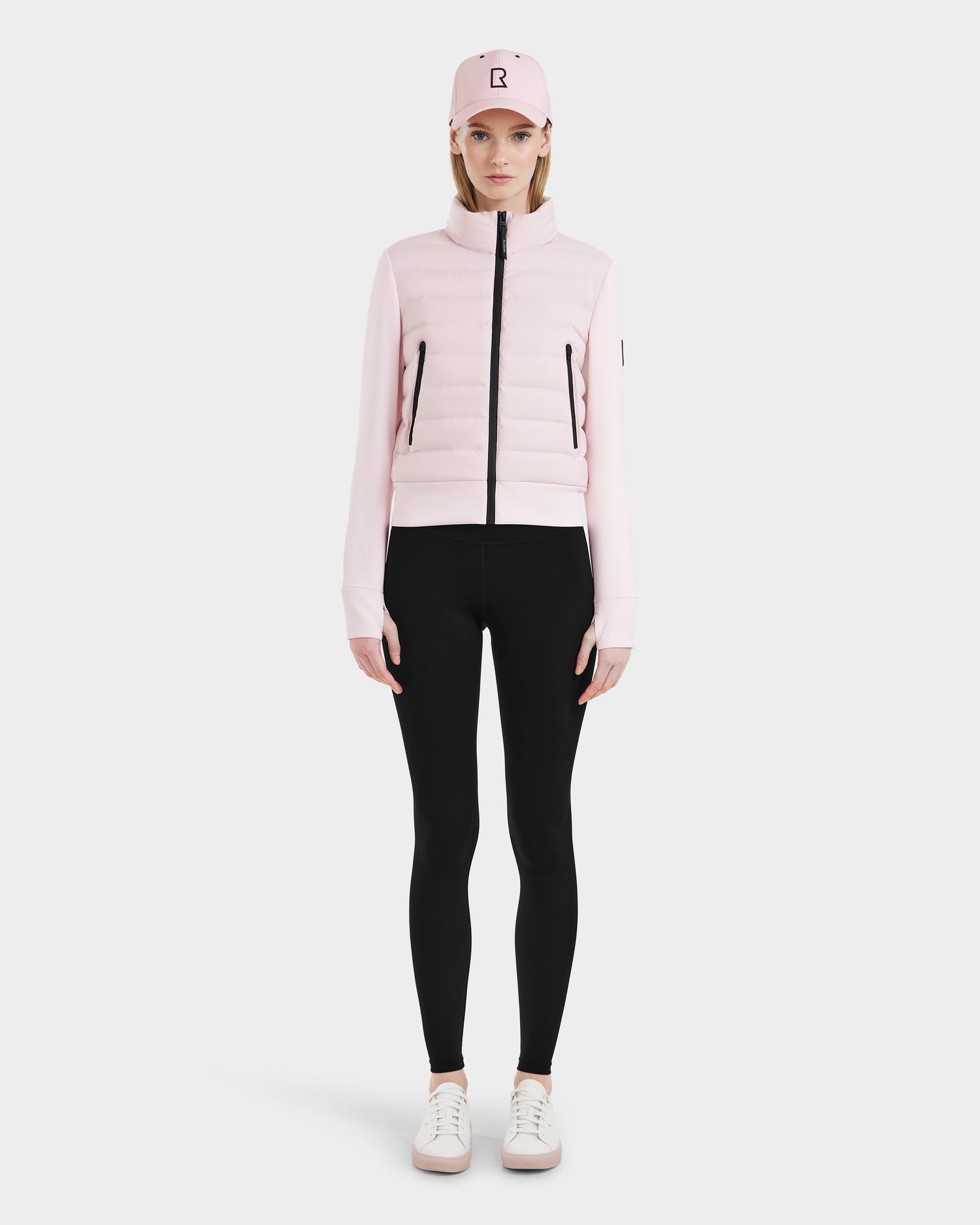 A woman faces forward against a white background, wearing the Rudsak CASSA - 8125057 light pink jacket, a matching pink cap, black leggings, and white sneakers.