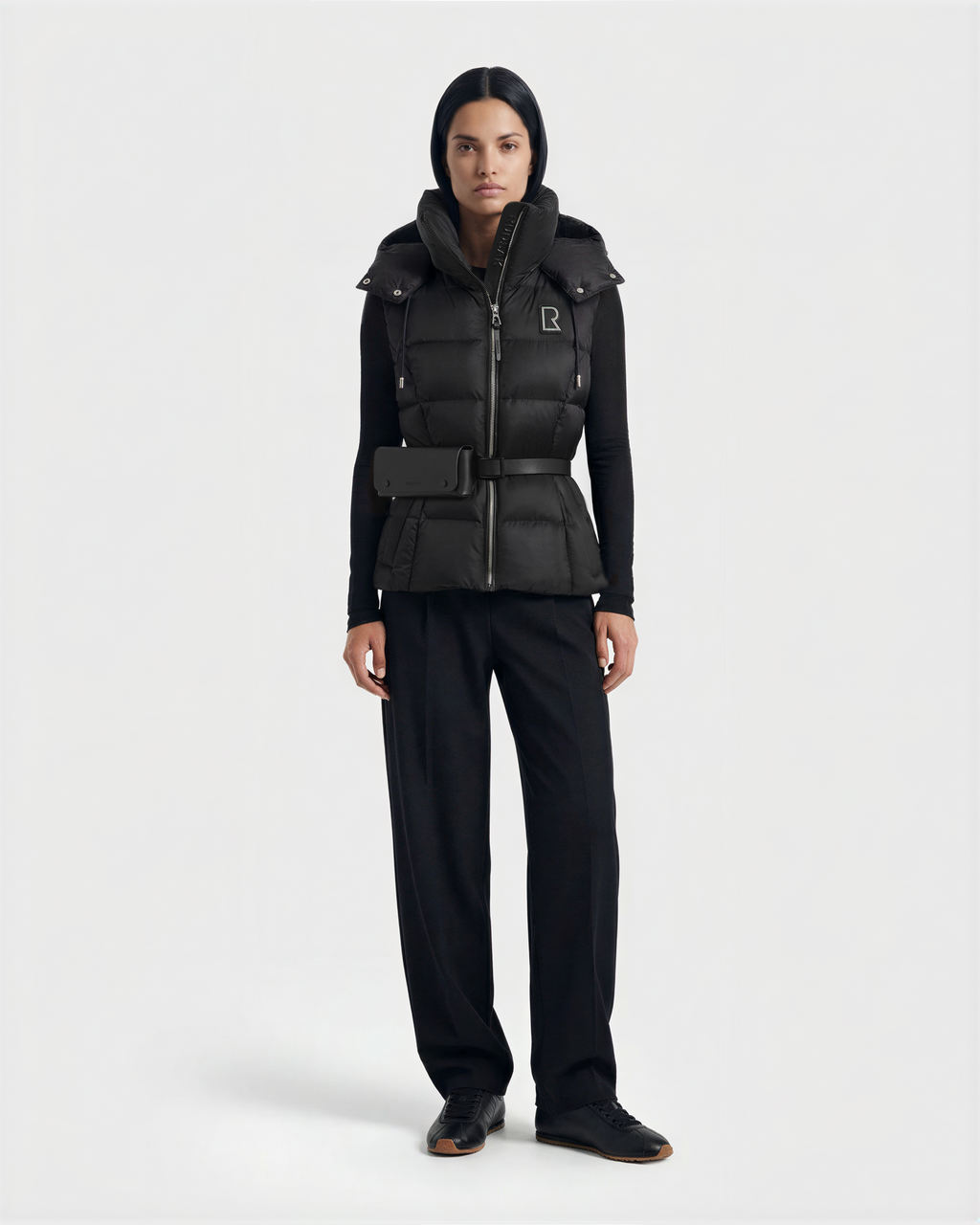 A woman with straight dark hair and a neutral expression faces forward, wearing the Rudsak CASSIDY N - 8125677 Black fitted down vest over a black long-sleeve top, wide-leg pants, and shoes against a plain light background.
