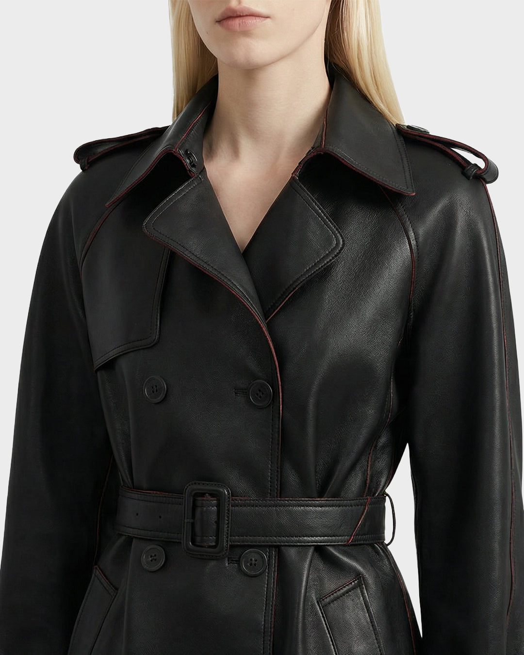 A woman with straight blonde hair wears the Rudsak CASSIE L-B - 8125553, a black bonded leather women’s trench coat with a belted waist, notched collar, button details, red piping accents, shoulder epaulets, and double-breasted design.