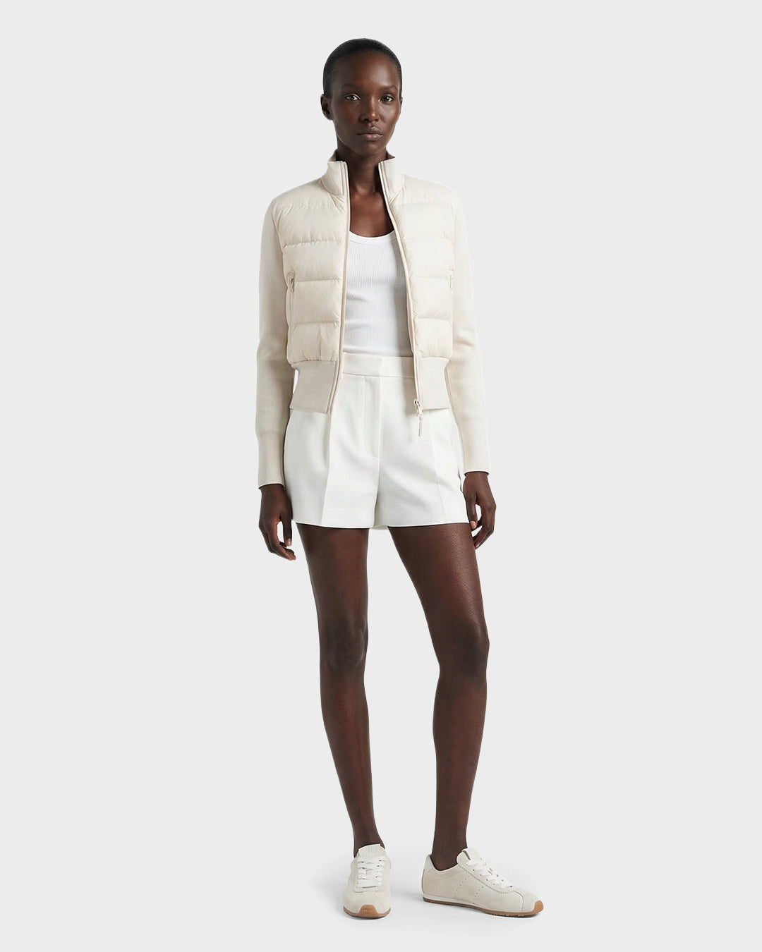 A woman faces forward wearing the Rudsak CLAREMONT - 8125502 Vanilla women's down jacket, styled with a white tank top, white shorts, and white sneakers against a plain light background.