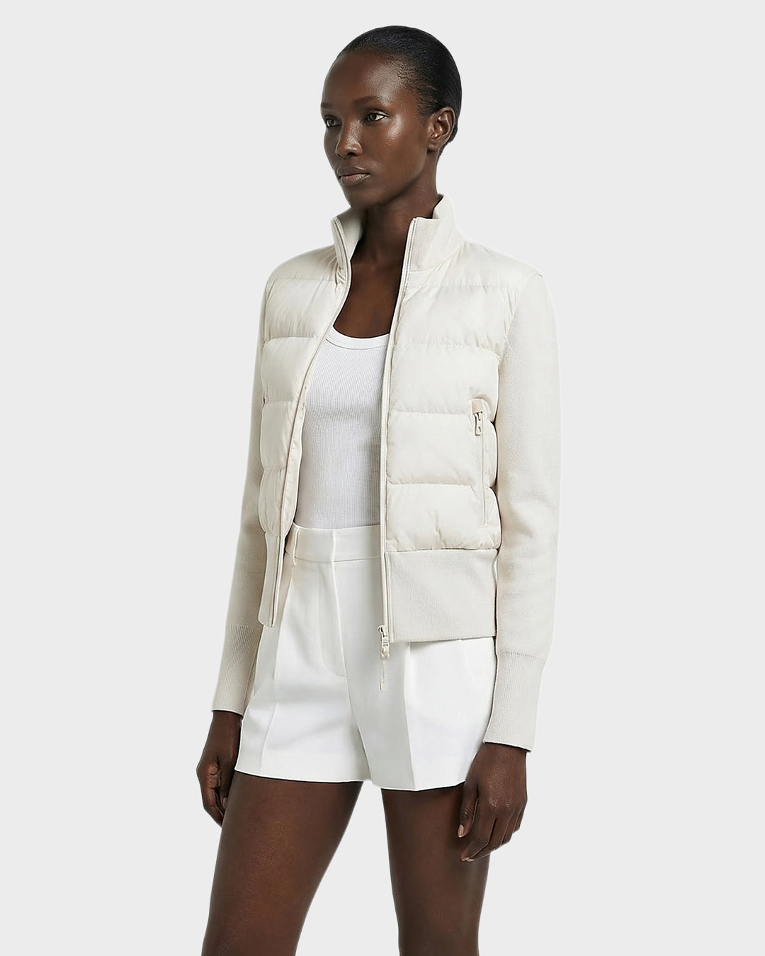 A woman in the Rudsak CLAREMONT - 8125502 Vanilla down jacket pairs it with a white tank top and shorts, posing against a plain background and embodying effortless style and lightweight warmth.