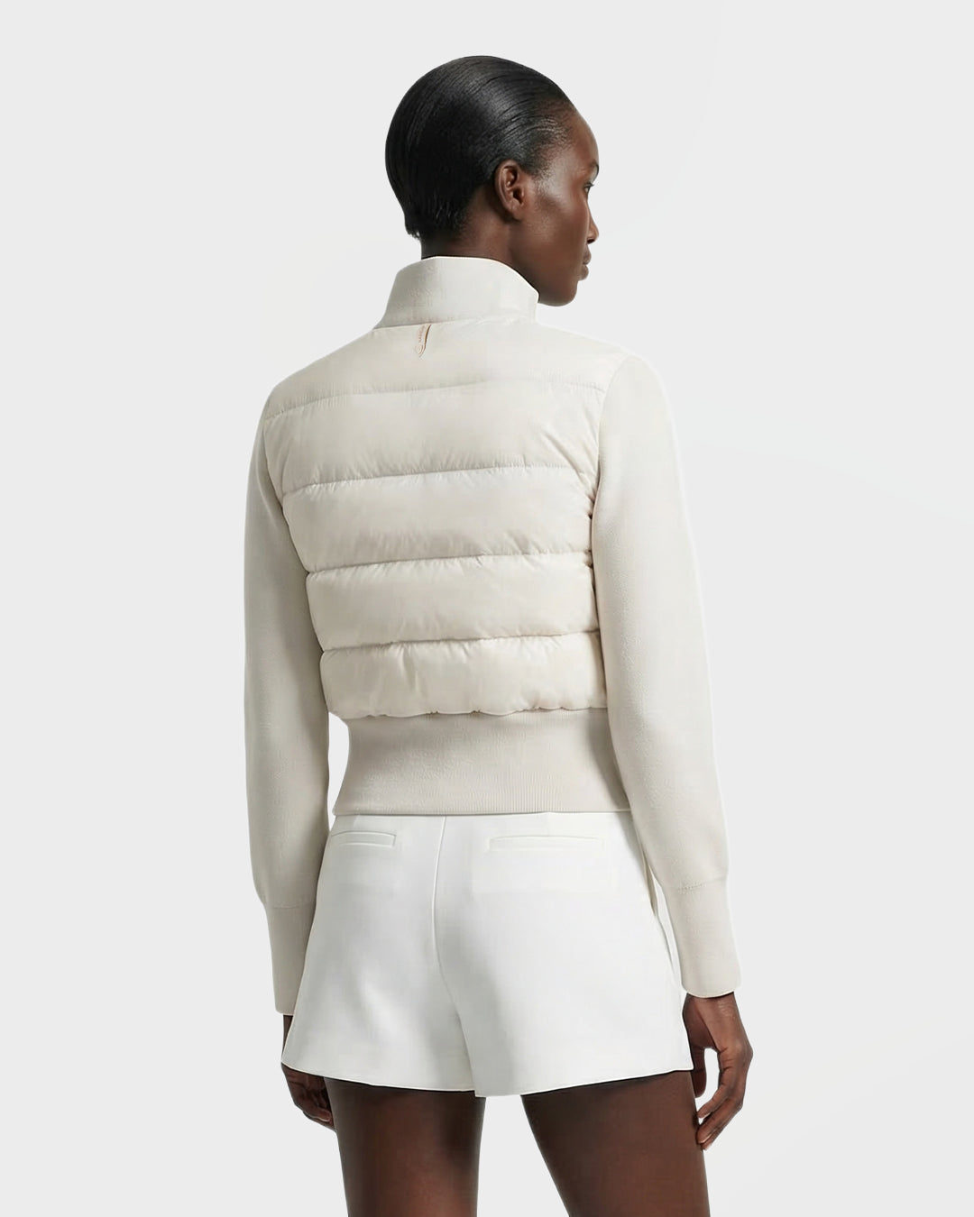 A woman with short, sleek hair is seen from the back wearing the Rudsak CLAREMONT - 8125502 Vanilla women’s down jacket and white shorts against a plain light background.