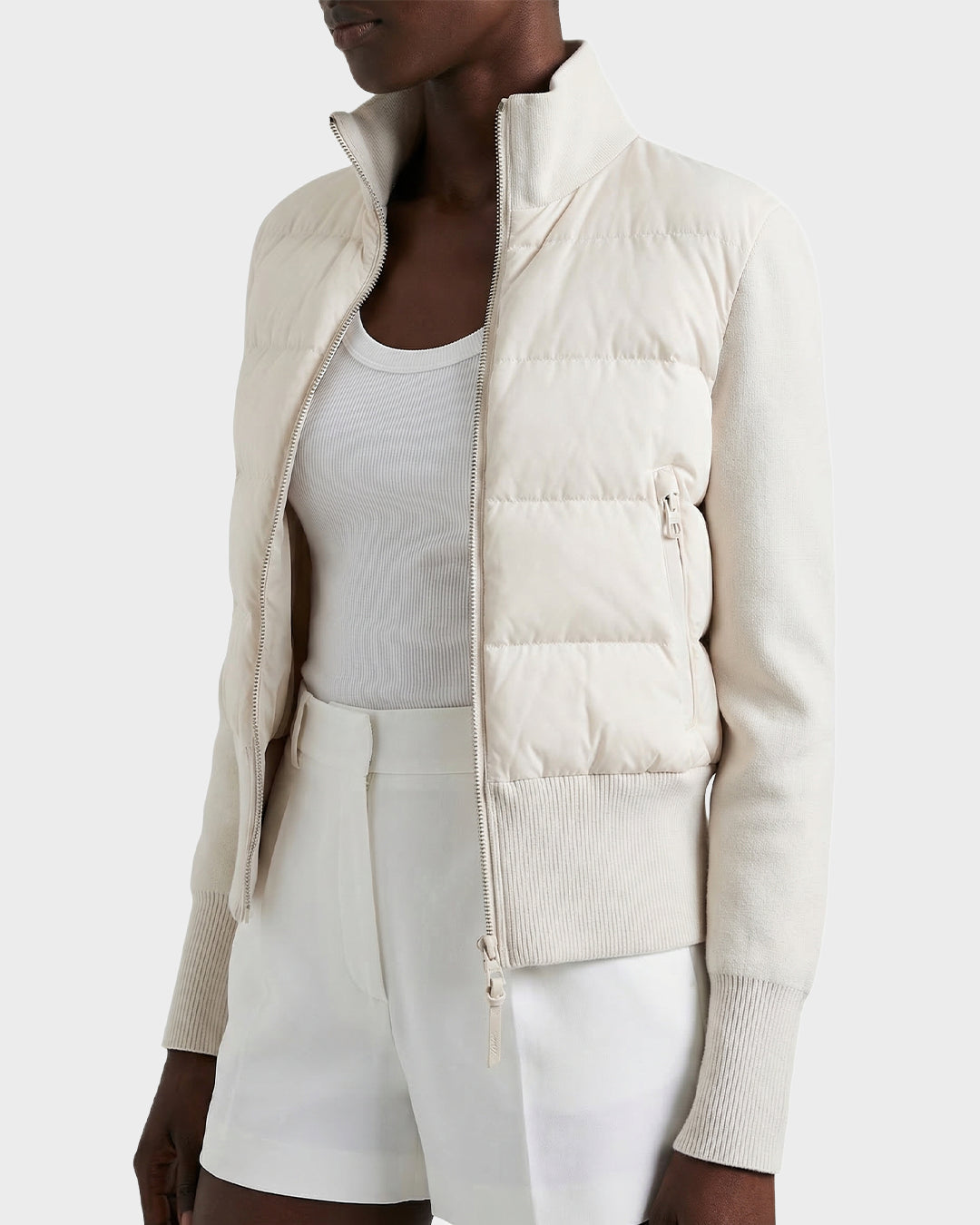 A person showcases effortless style and lightweight warmth in the Rudsak CLAREMONT - 8125502 Vanilla women’s down jacket, layered over a white tank top and high-waisted white shorts against a plain background.