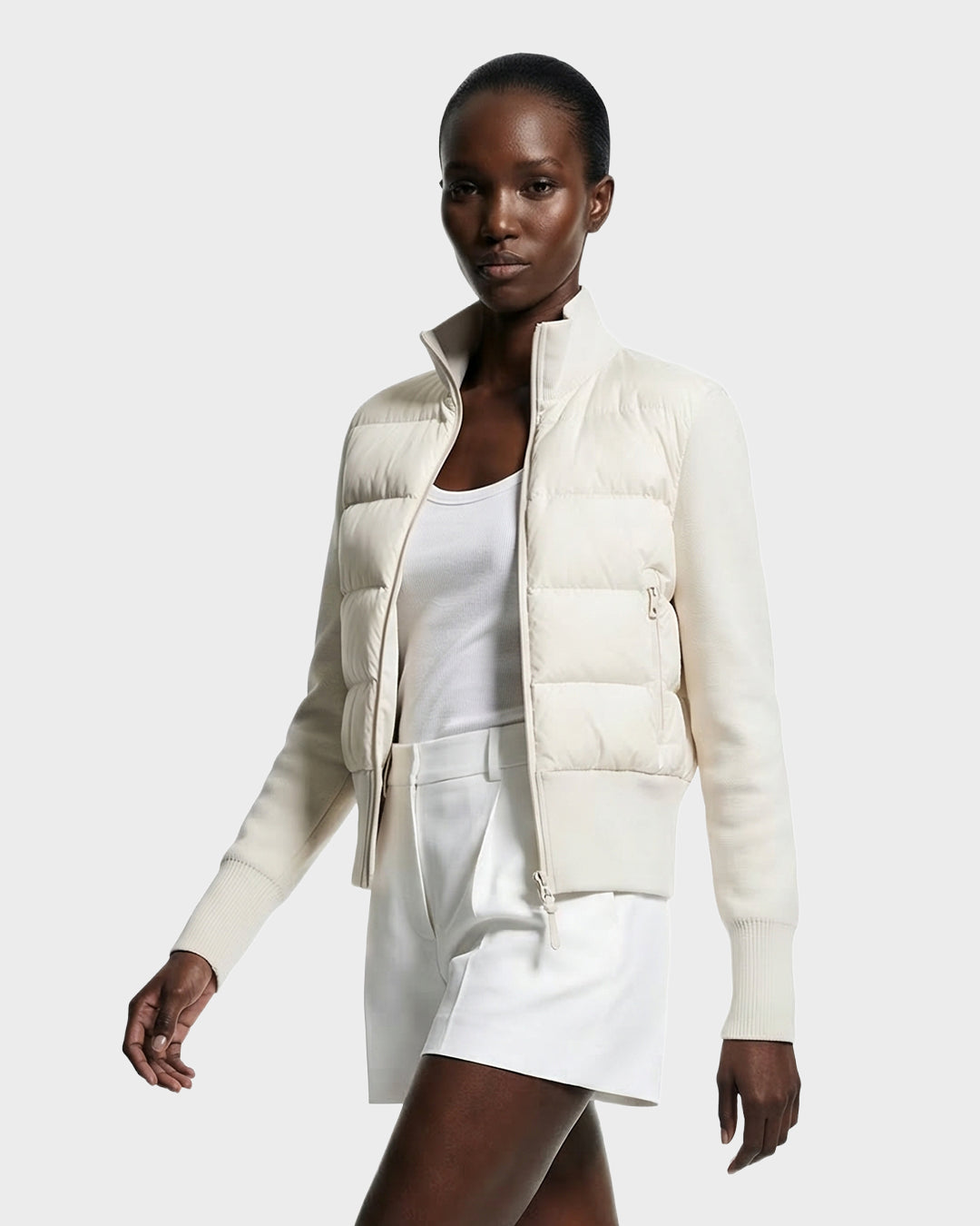 A woman in the Rudsak CLAREMONT - 8125502 Vanilla down jacket pairs it with a white tank top and shorts, walking with a neutral expression against a plain light background, highlighting its lightweight warmth and effortless style.