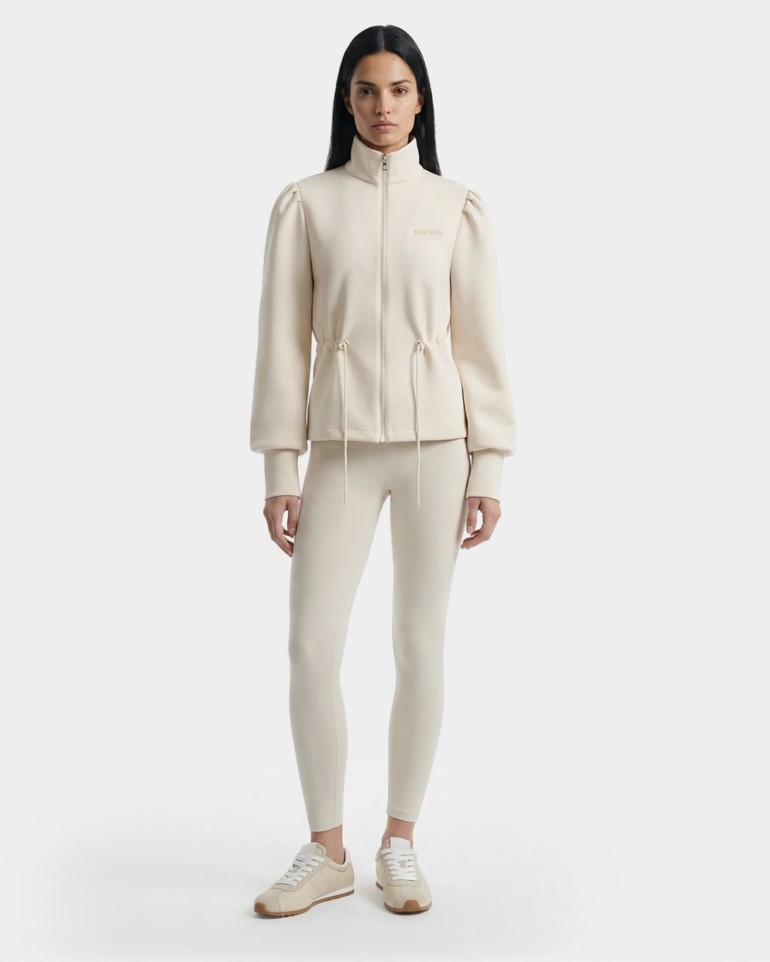 A woman faces forward in a sports-casual look, wearing the Rudsak ELODIE - 8526039 Iceland light beige zip-up sweater jacket with drawstrings, matching leggings, and white sneakers. Her long dark hair stands out against a minimalist white background.