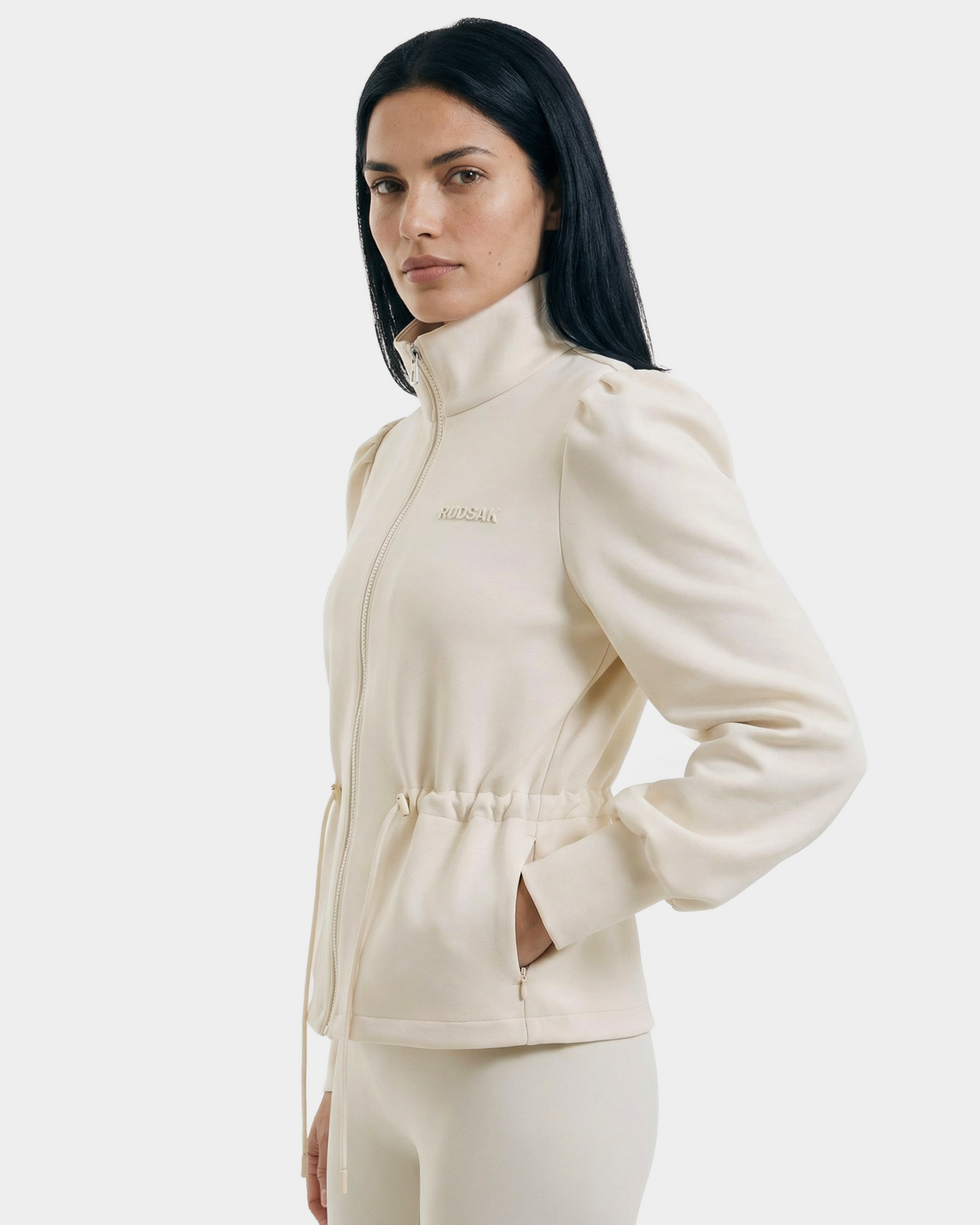 A woman with long, straight dark hair wears the Rudsak ELODIE - 8526039 Iceland zip-up sweater jacket with high collar and side pockets, paired with matching pants, standing facing left against a plain light background.