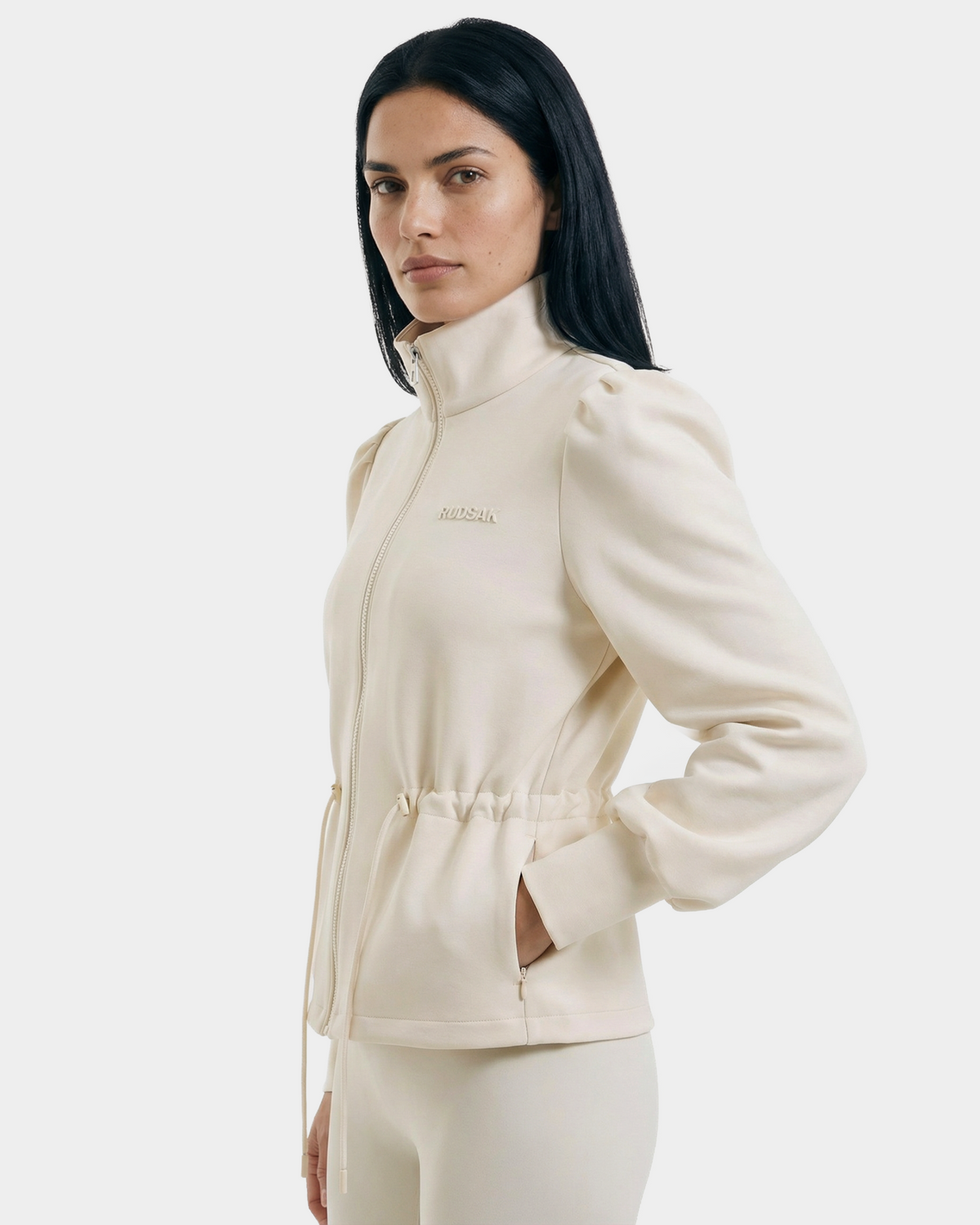A woman with long, straight dark hair wears the Rudsak ELODIE - 8526039 Iceland zip-up sweater jacket with high collar and side pockets, paired with matching pants, standing facing left against a plain light background.