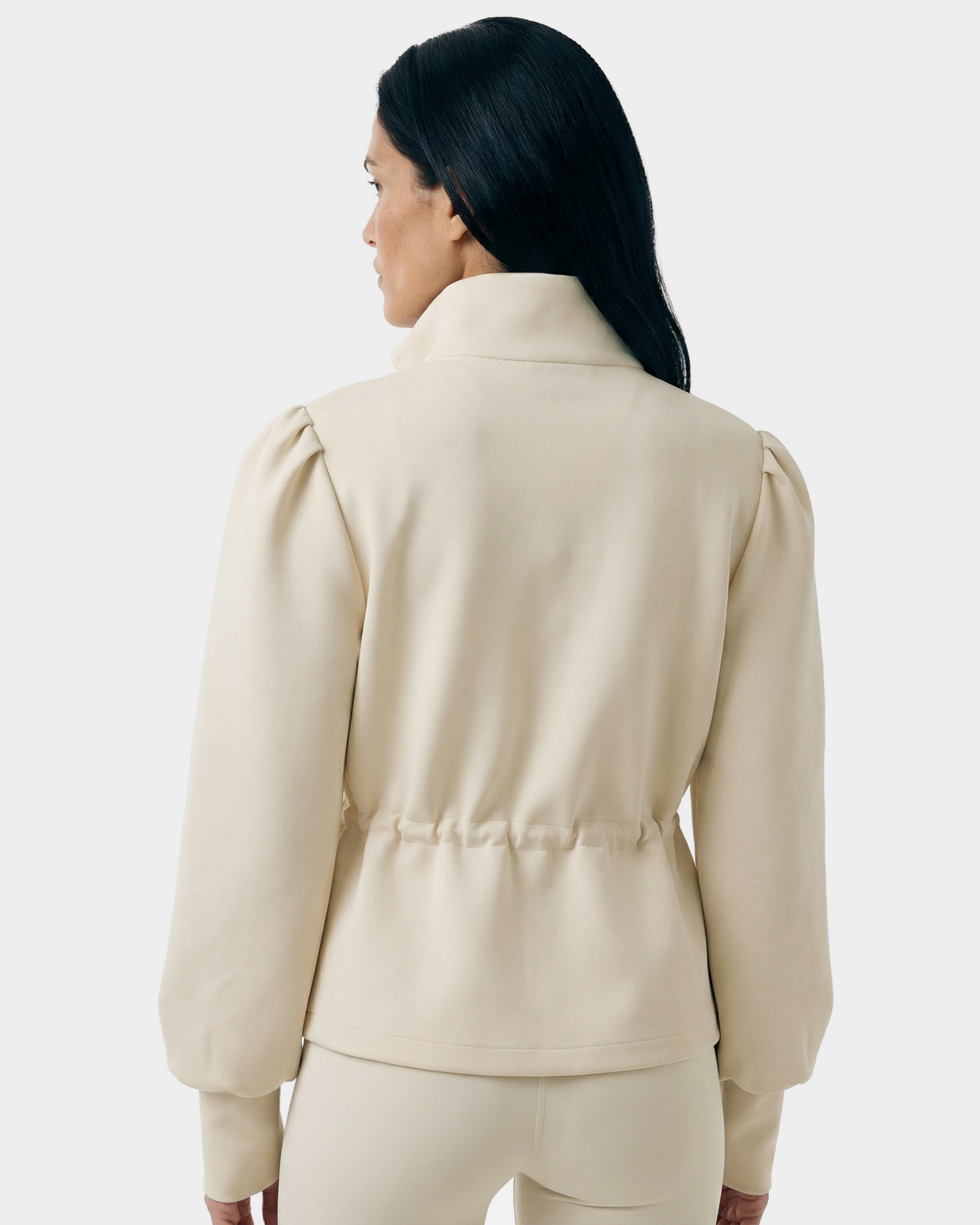 A woman with long black hair faces away, wearing the Rudsak ELODIE - 8526039 Iceland zip-up sweater jacket and matching pants. The high collar and gathered waist stand out against the plain light grey background.