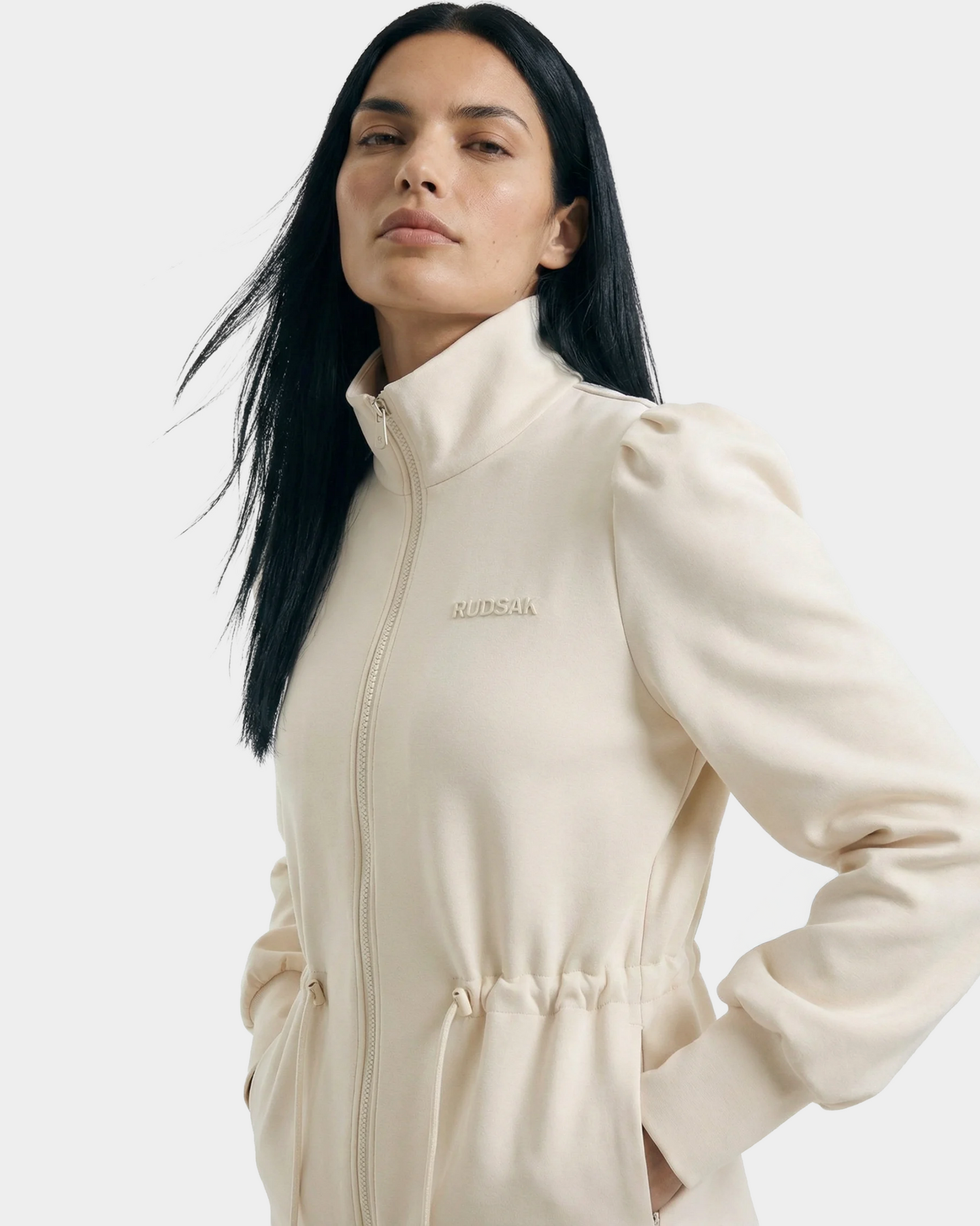 A woman with long black hair wears the Rudsak ELODIE - 8526039 Iceland cream zip-up sweater jacket with a gathered waist and high collar, hands in pockets. The lightweight layer pops against a plain light background.