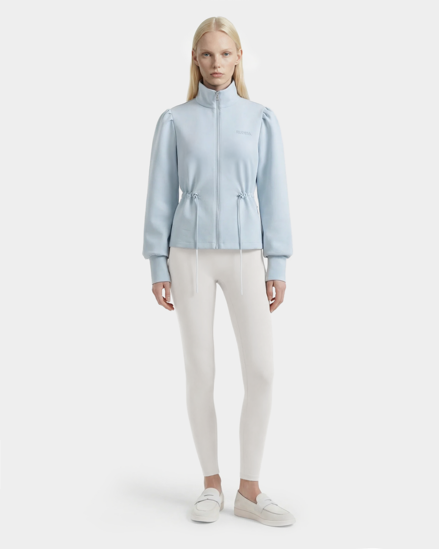 A woman with long blonde hair wears the Rudsak ELODIE - 8526039 Powder Blue zip-up sweater jacket, paired with white leggings and slip-on shoes, standing against a plain light background.