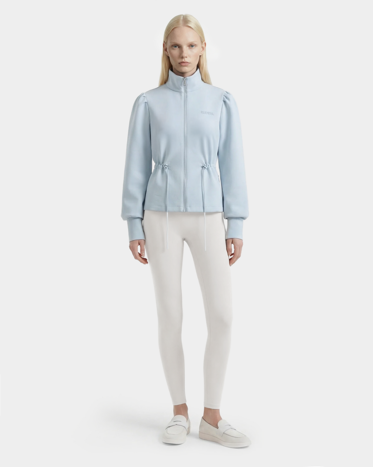 A woman with long blonde hair wears the Rudsak ELODIE - 8526039 Powder Blue zip-up sweater jacket, paired with white leggings and slip-on shoes, standing against a plain light background.