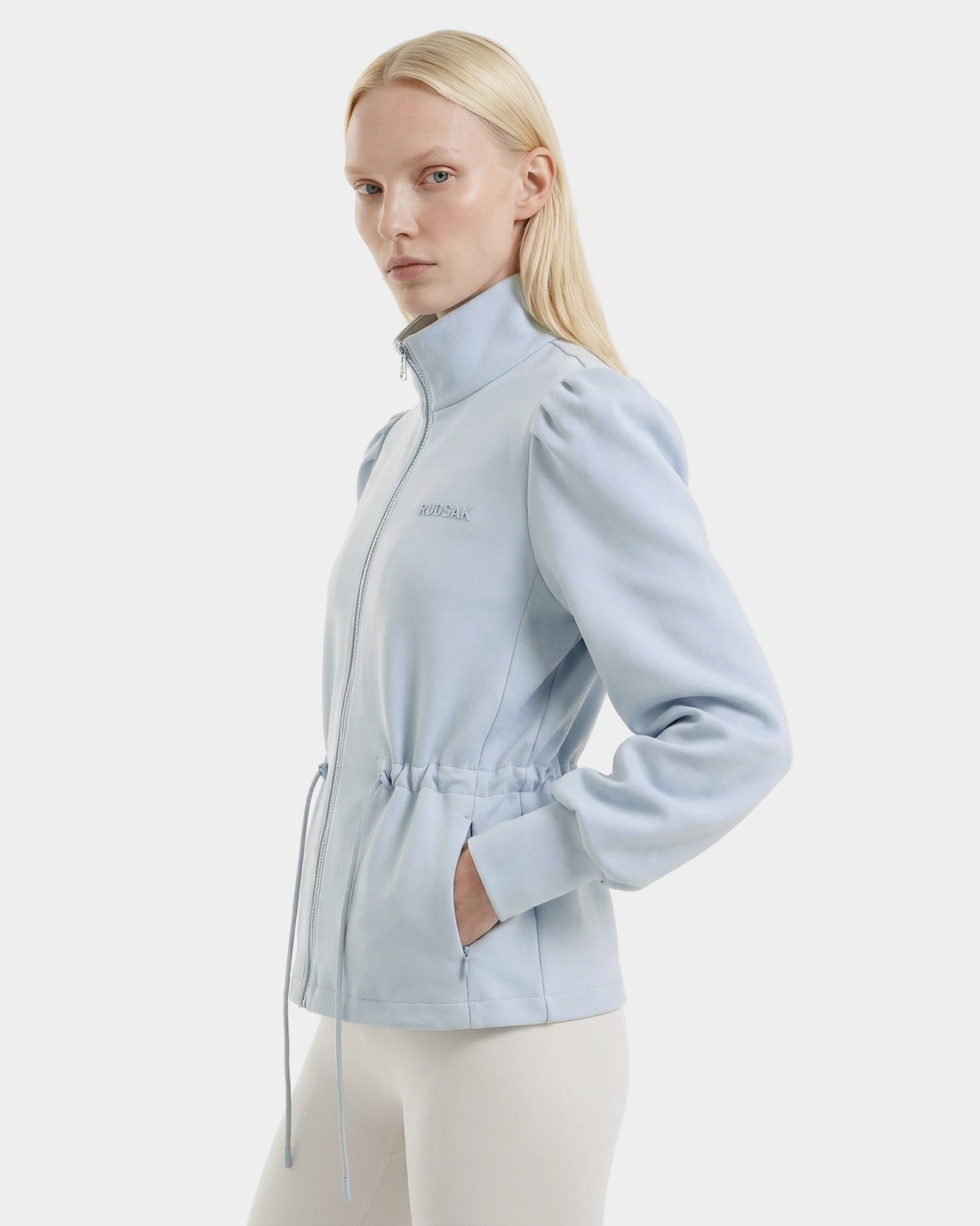 A woman with straight blonde hair wears the Rudsak ELODIE - 8526039 Powder Blue zip-up sweater jacket, featuring a high collar and drawstring waist, paired with white pants against a plain white background.