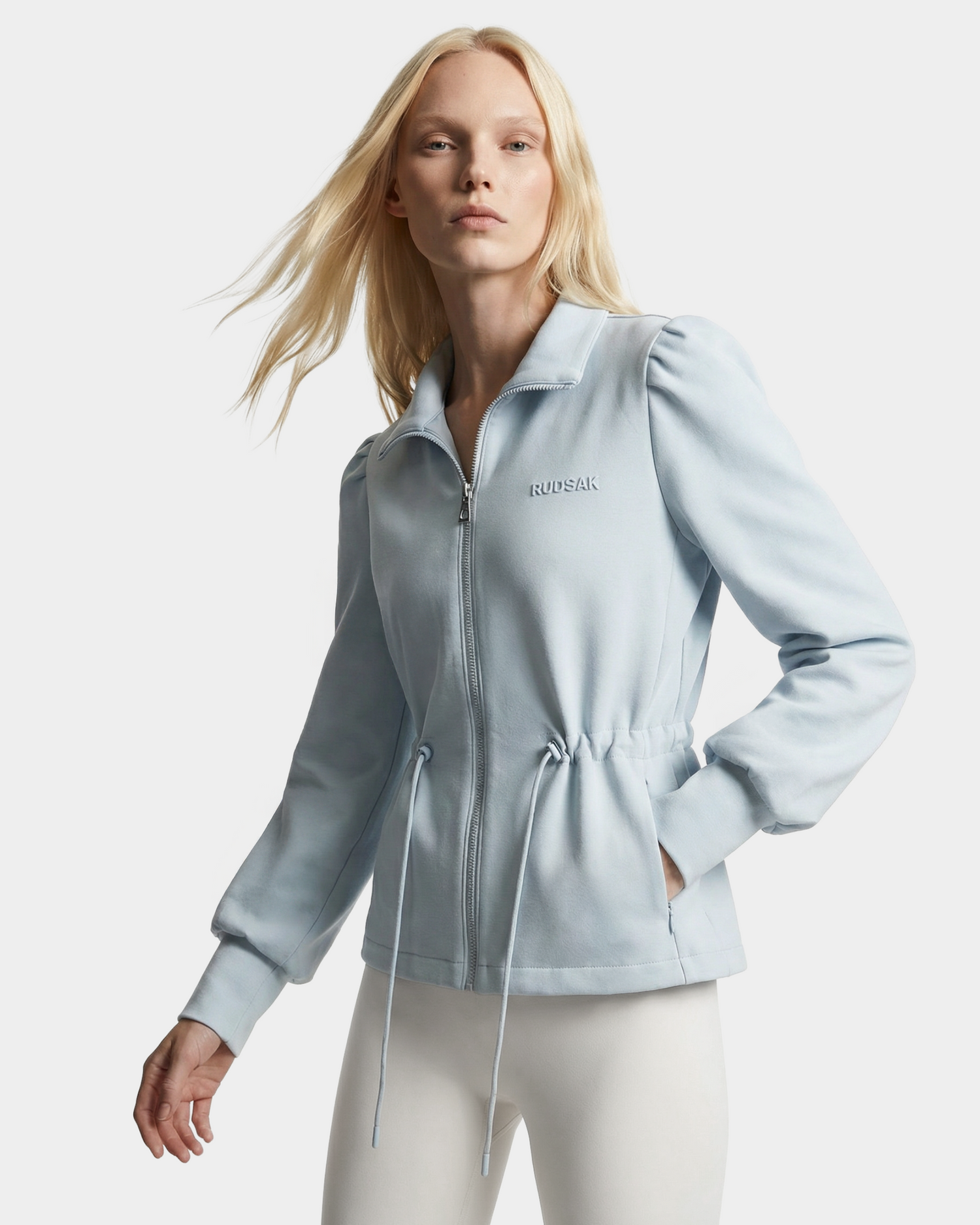 A woman with long blonde hair models the Rudsak ELODIE - 8526039 Powder Blue zip-up sweater jacket with a drawstring waist and puffed sleeves, paired with white pants, against a plain white background.