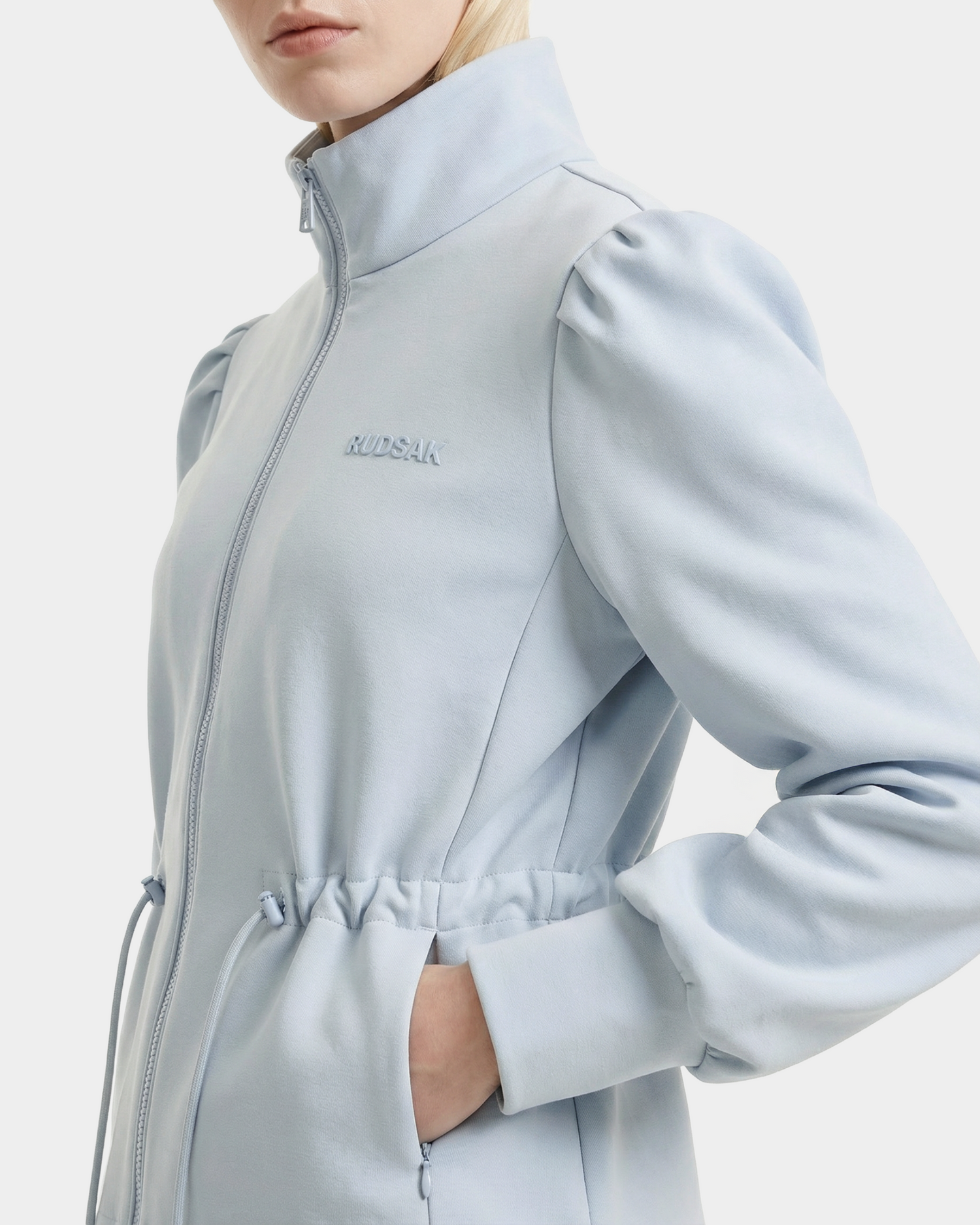 A woman wears the Rudsak ELODIE - 8526039 Powder Blue zip-up sweater jacket featuring puffed shoulders, a high collar, elastic drawstring waist, and embroidered branding on the chest—a perfect lightweight sports-casual layer.