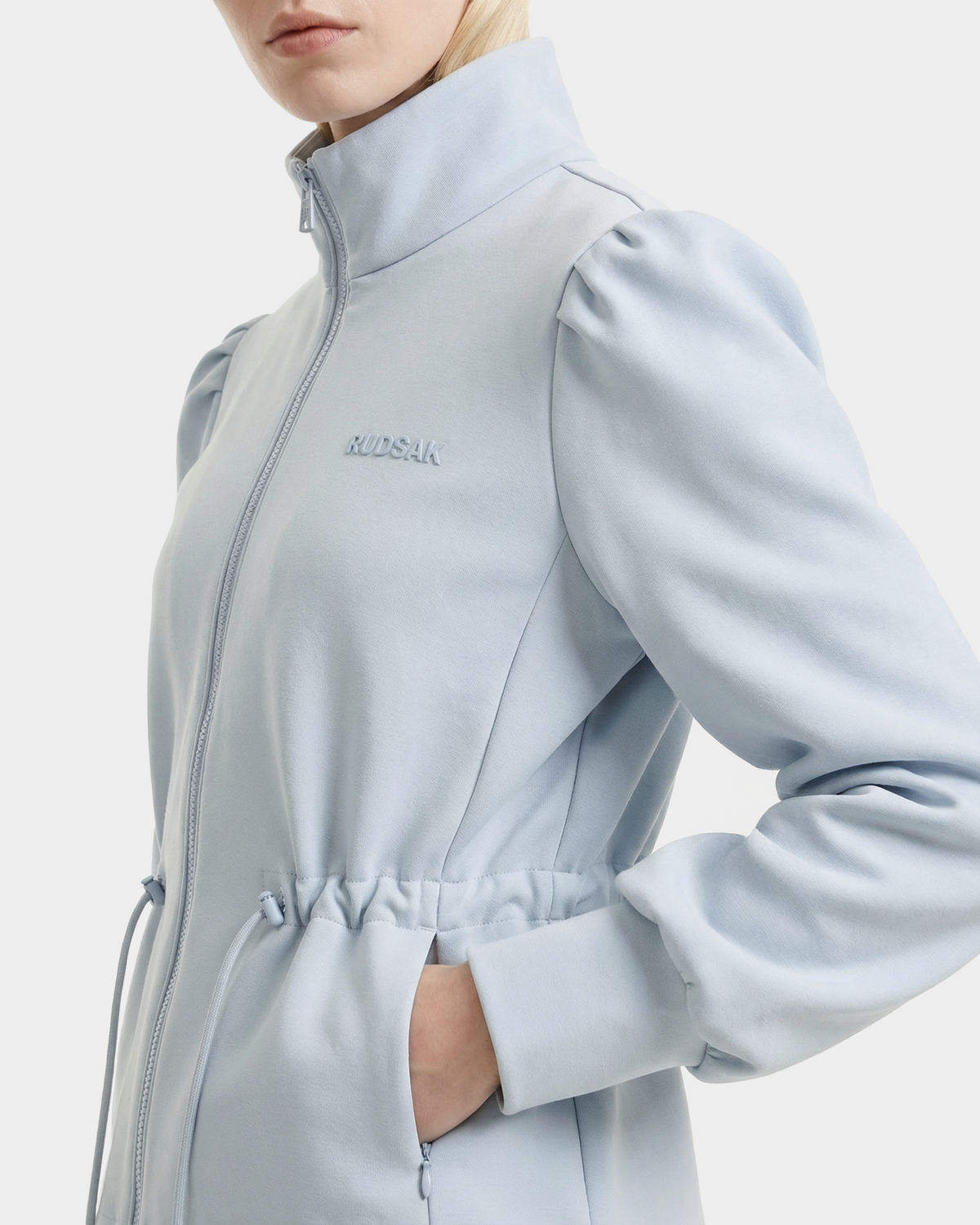 A woman wears the Rudsak ELODIE - 8526039 Powder Blue zip-up sweater jacket featuring puffed shoulders, a high collar, elastic drawstring waist, and embroidered branding on the chest—a perfect lightweight sports-casual layer.