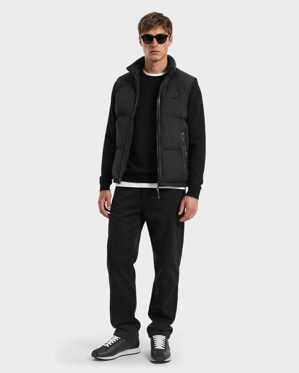 A man models the Rudsak FREDDY - 6125571 Black men’s puffer vest over a black long-sleeve shirt, paired with black pants and sunglasses, standing against a light gray background to showcase stylish lightweight layering.