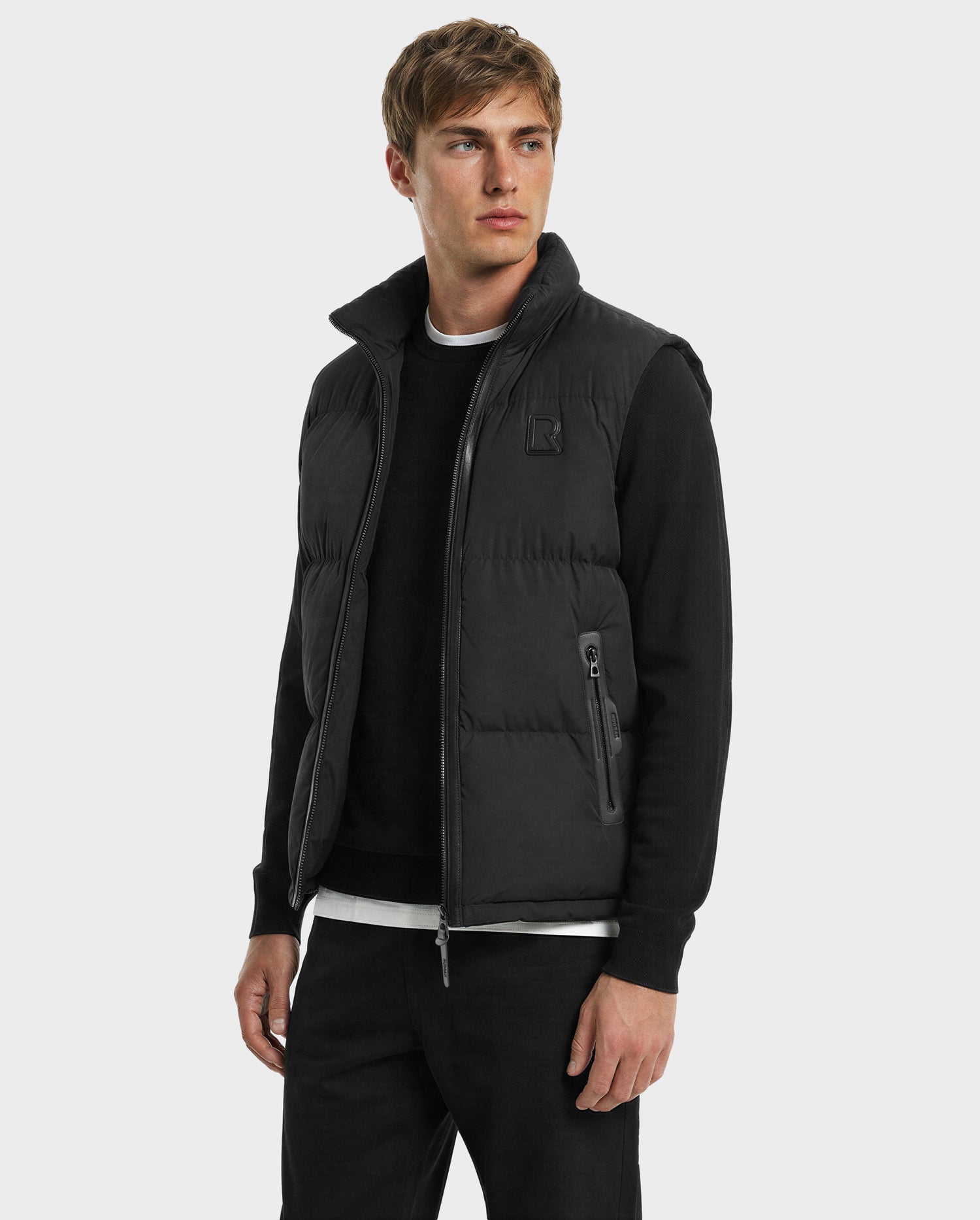 A young man models the Rudsak FREDDY - 6125571 Black men’s puffer vest, featuring premium down and lightweight warmth, layered over a black long-sleeve shirt and matching pants, against a plain light gray background.