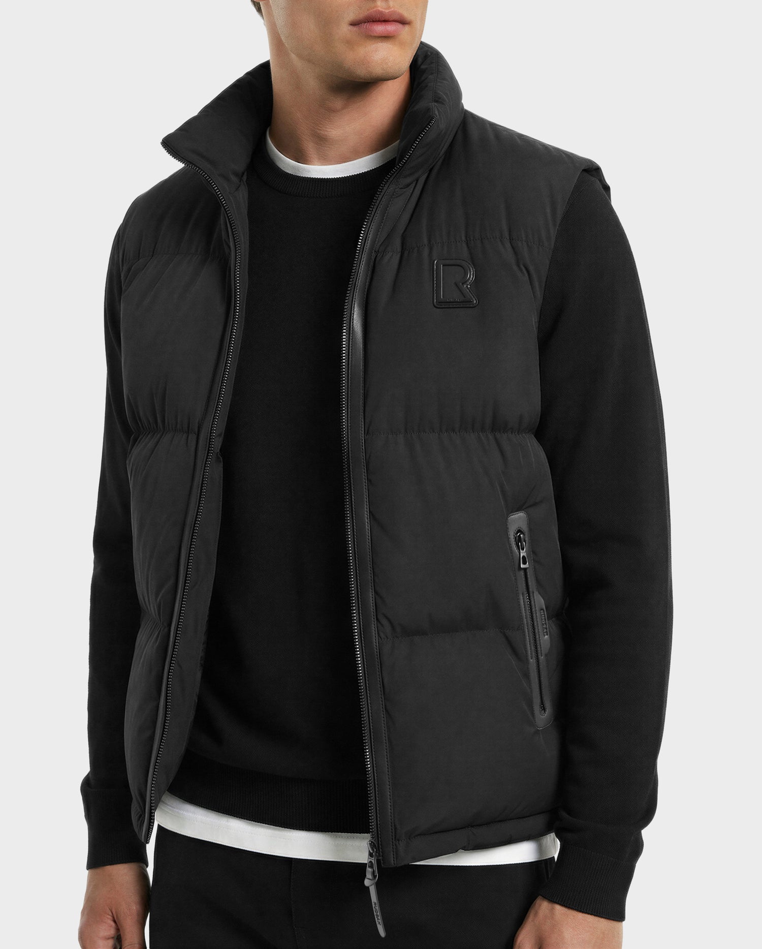 A man wears Rudsak’s FREDDY - 6125571 Black men’s puffer vest over a black crew neck sweatshirt and white t-shirt, with his left hand in his pocket, showcasing premium down layering against a plain light gray background.
