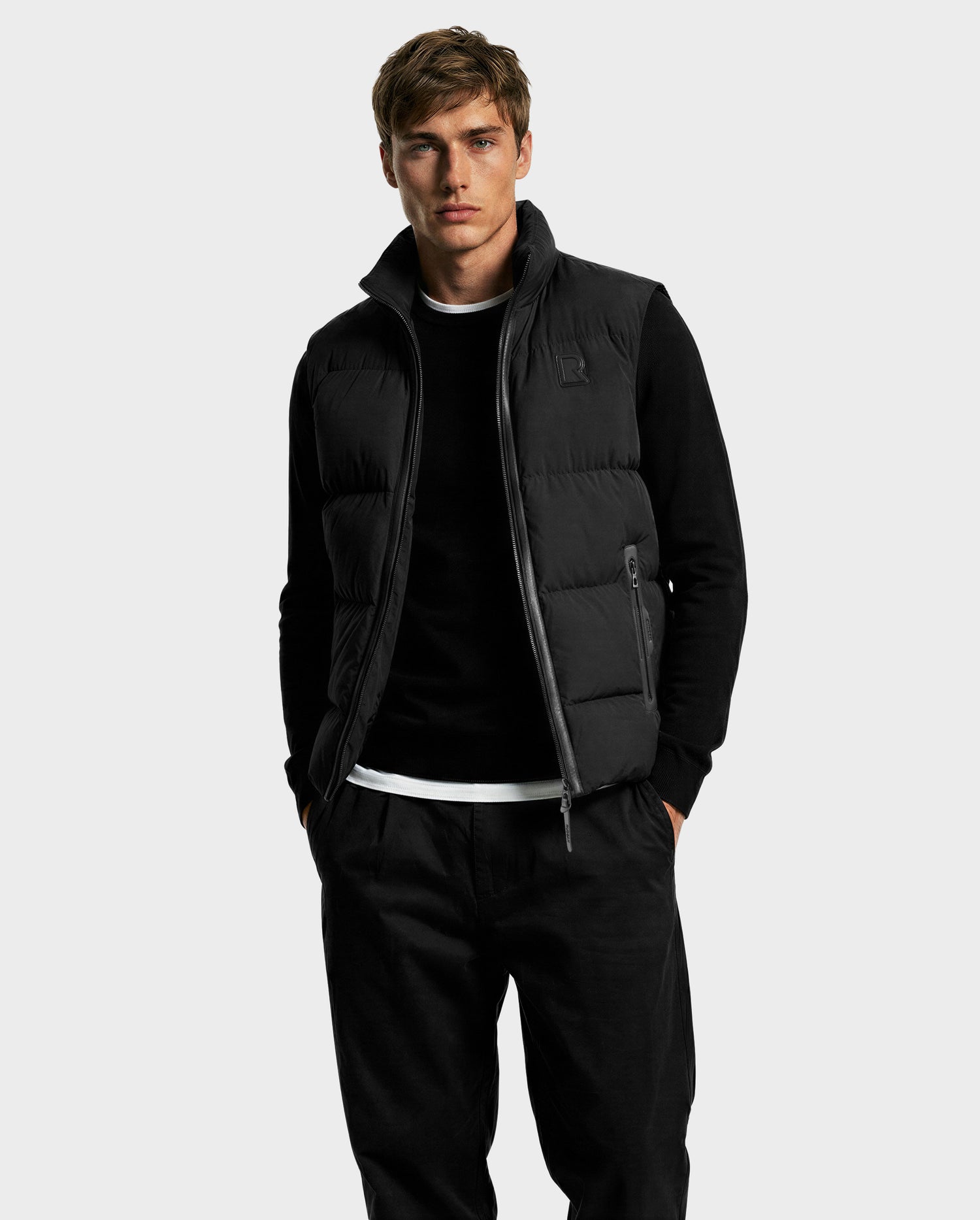 A man wears the Rudsak FREDDY - 6125571 Black premium down puffer vest over a black long-sleeve shirt and black pants, standing with hands in pockets to showcase effortless lightweight layering against a plain light gray background.