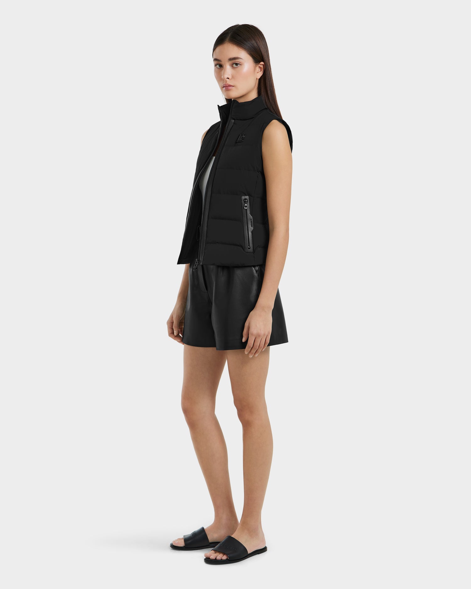 A woman with straight brown hair and a neutral look stands against a plain background, wearing the Rudsak GABRIELA - 8125014 Black women’s down puffer vest with vegan leather trim over a black outfit and slide sandals.
