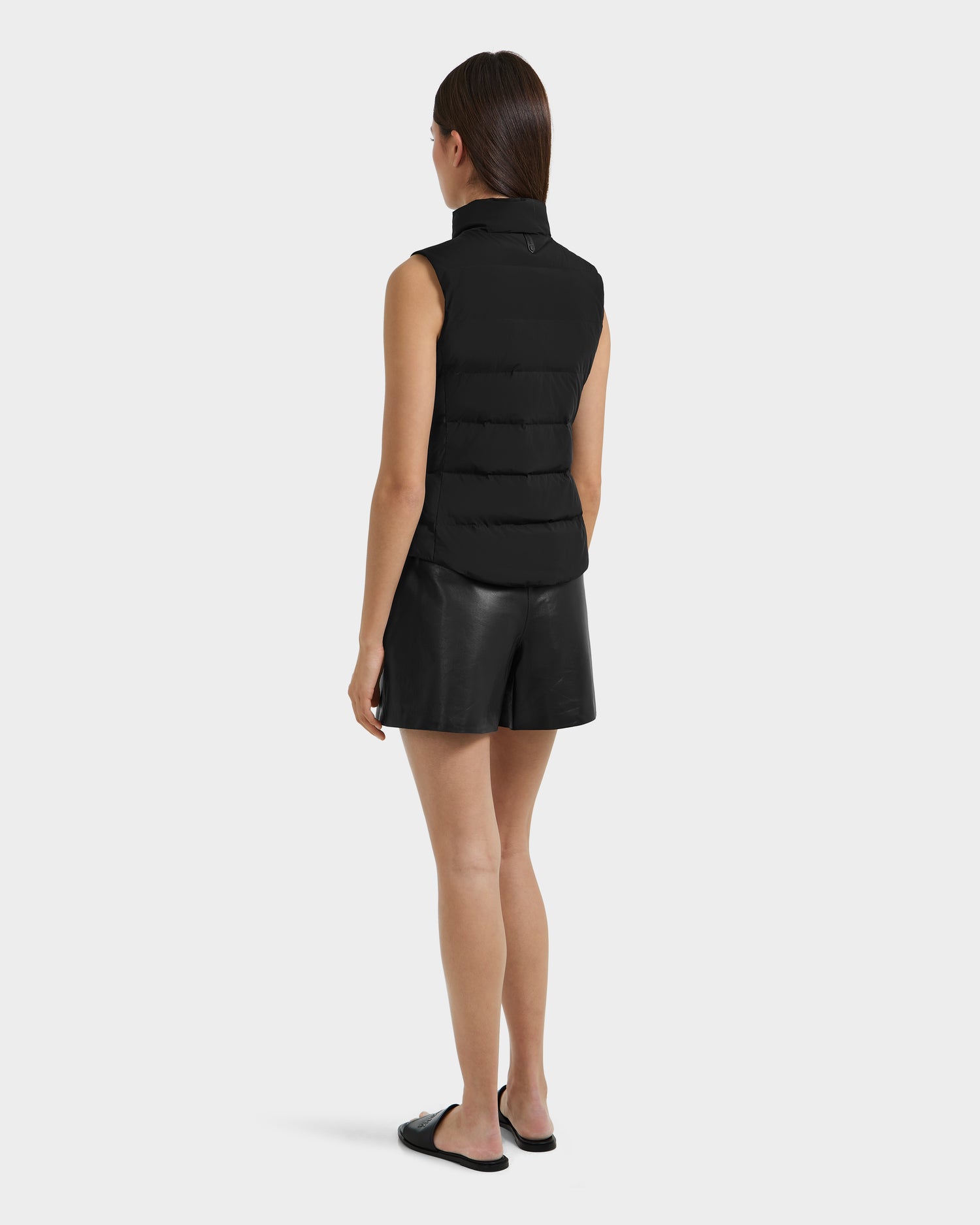 A woman faces away, wearing the Rudsak GABRIELA - 8125014 Black women’s down puffer vest with vegan leather trim, black leather shorts, and black slide sandals against a plain white background.