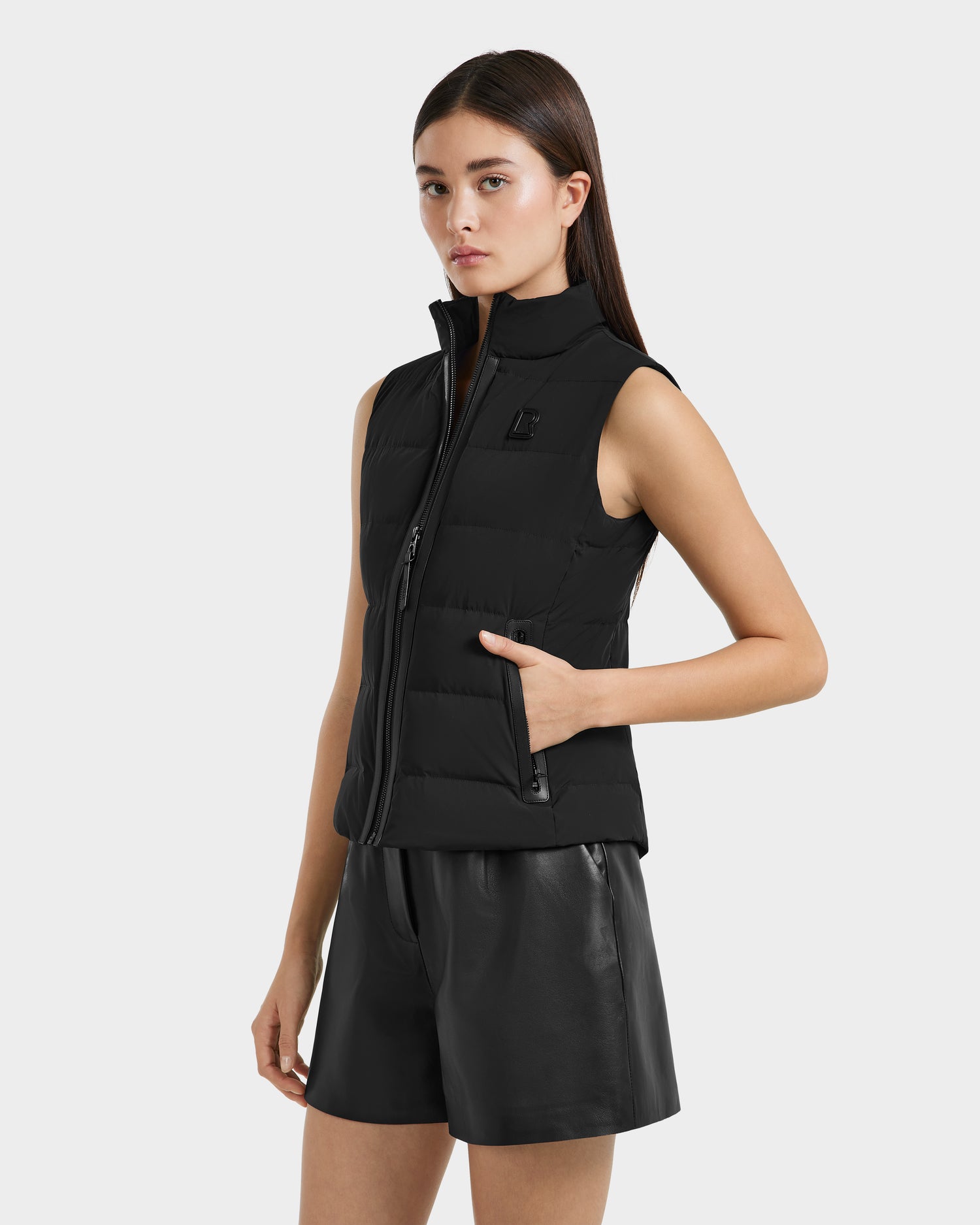 A woman with straight brown hair wears the Rudsak GABRIELA - 8125014 Black lightweight puffer vest with vegan leather trim and black leather shorts, standing with one hand in her vest pocket against a plain light background.