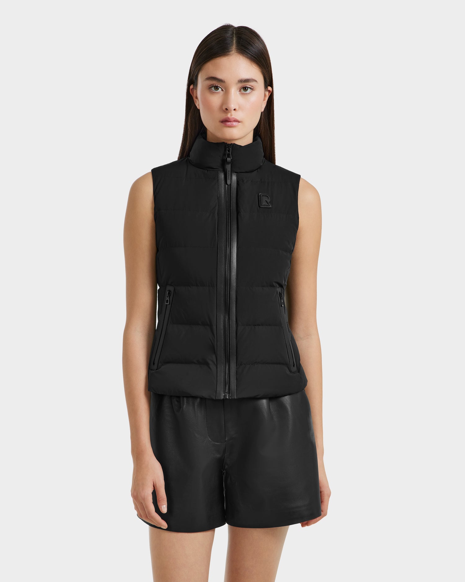 A woman with straight brown hair wears the Rudsak GABRIELA - 8125014 Black lightweight puffer vest with vegan leather trim and black shorts, standing against a plain light background.