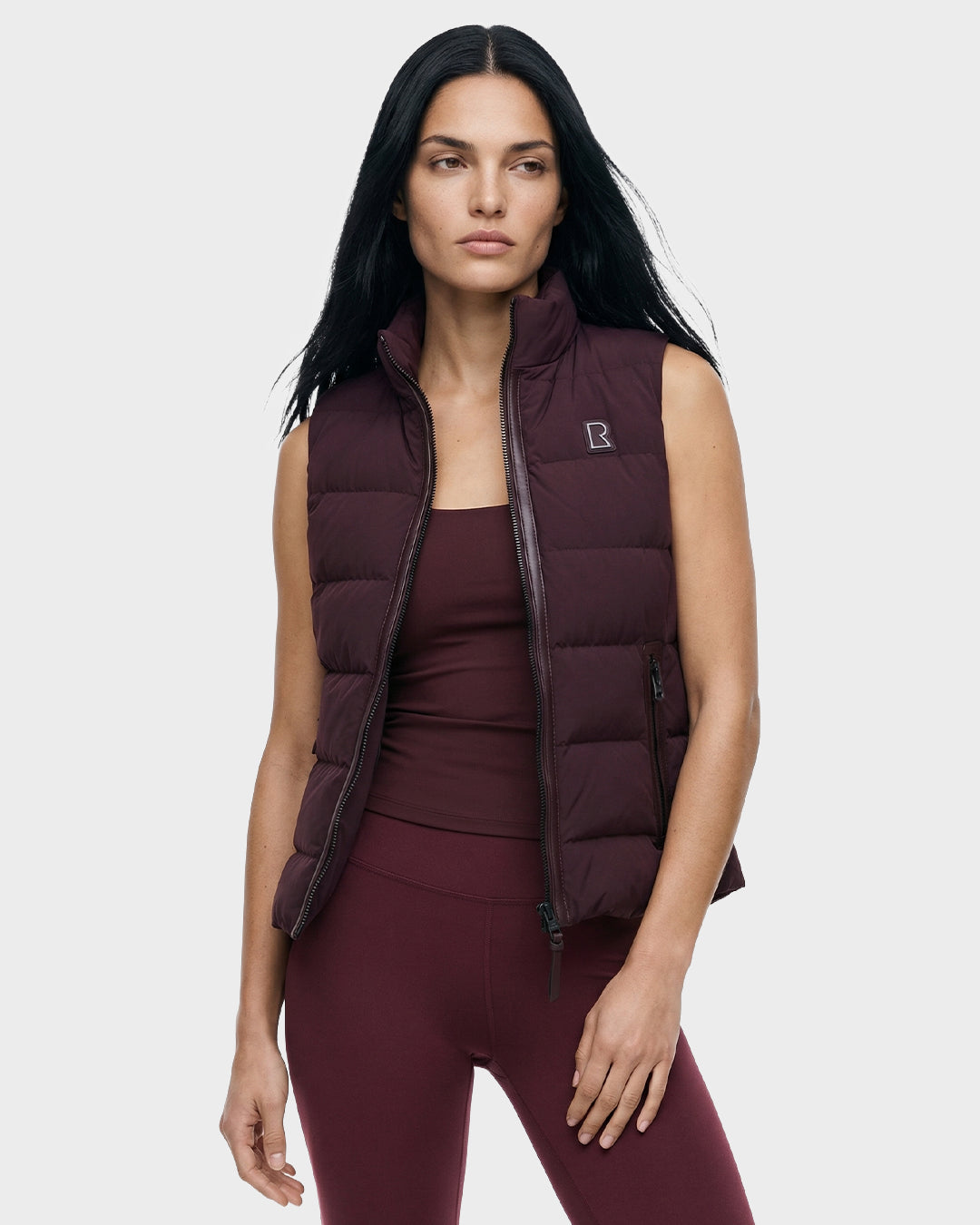 A woman with long dark hair models the Rudsak GABRIELA - 8125014 Bordeaux women’s down puffer vest over a matching tank top and leggings, standing against a plain light gray background.