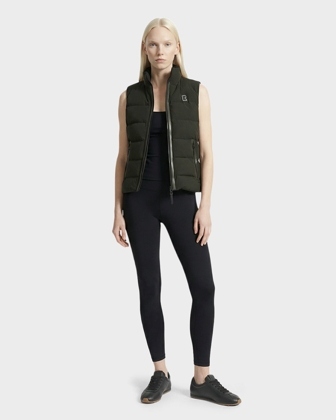 A woman with long blonde hair models the Rudsak GABRIELA - 8125014 Dark Olive lightweight puffer vest with vegan leather trims over an all-black outfit, standing against a plain light gray background.
