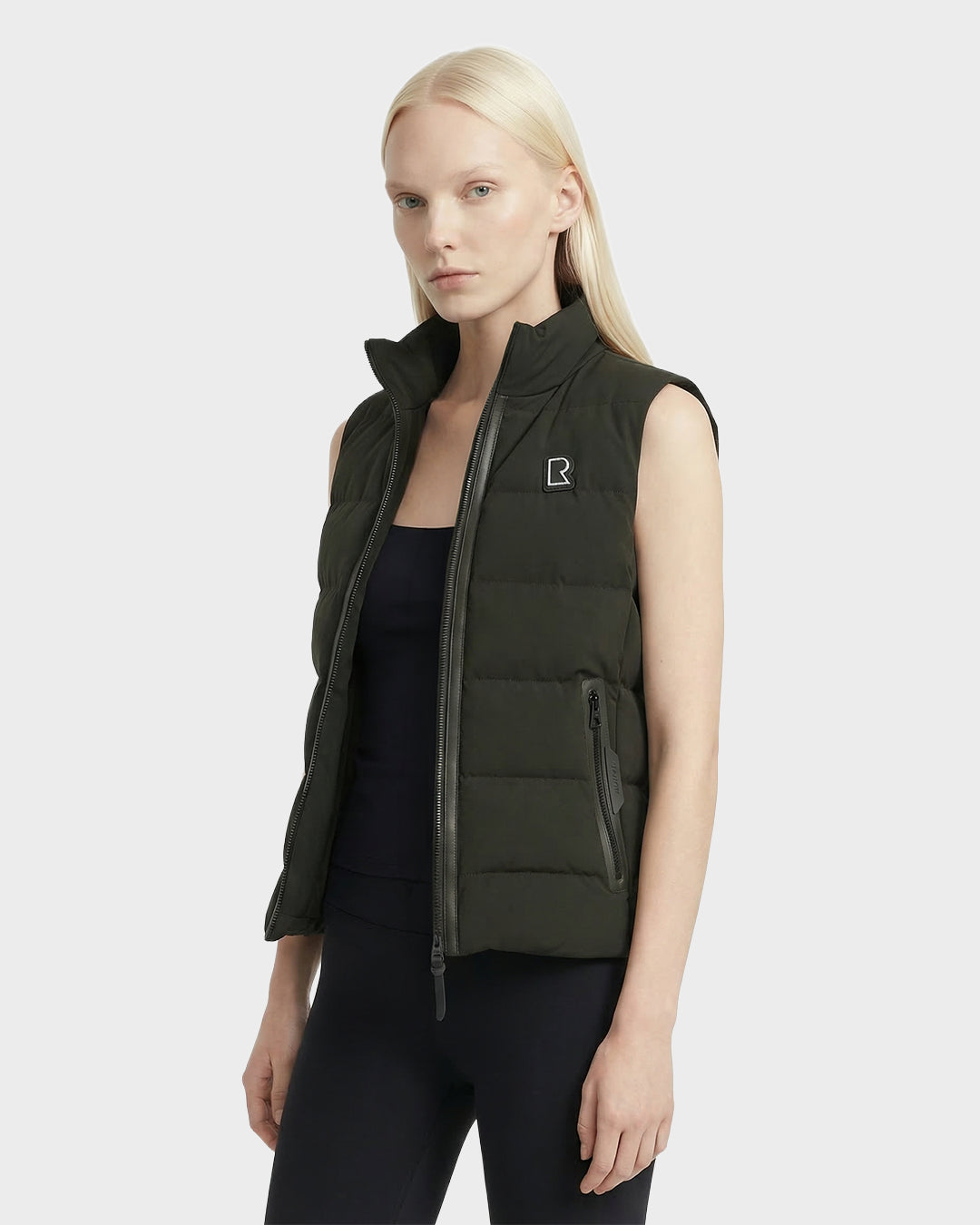 A woman with straight blonde hair wears the Rudsak GABRIELA - 8125014 Dark Olive lightweight puffer vest with vegan leather trims and a small R logo over a black top and leggings, standing out against a plain light gray background.