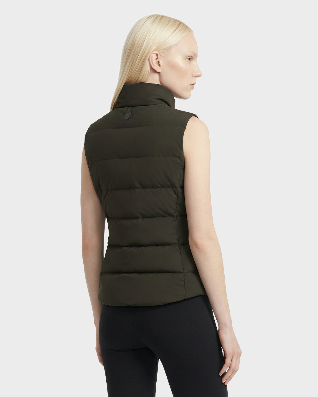A person with long blonde hair wears the Rudsak GABRIELA - 8125014 Dark Olive women's down puffer vest and black leggings, standing with their back turned slightly to the side against a plain light background.