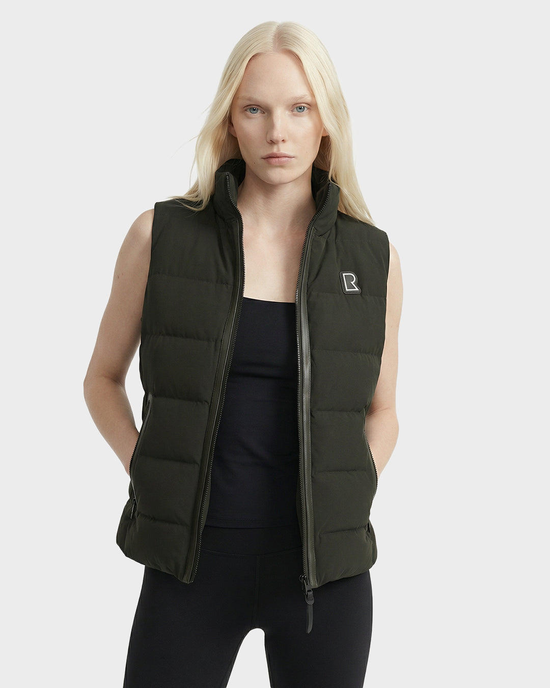 A woman with long blonde hair models the Rudsak GABRIELA - 8125014 Dark Olive lightweight puffer vest with vegan leather trims over a black top and leggings, standing with hands in vest pockets against a plain light background.