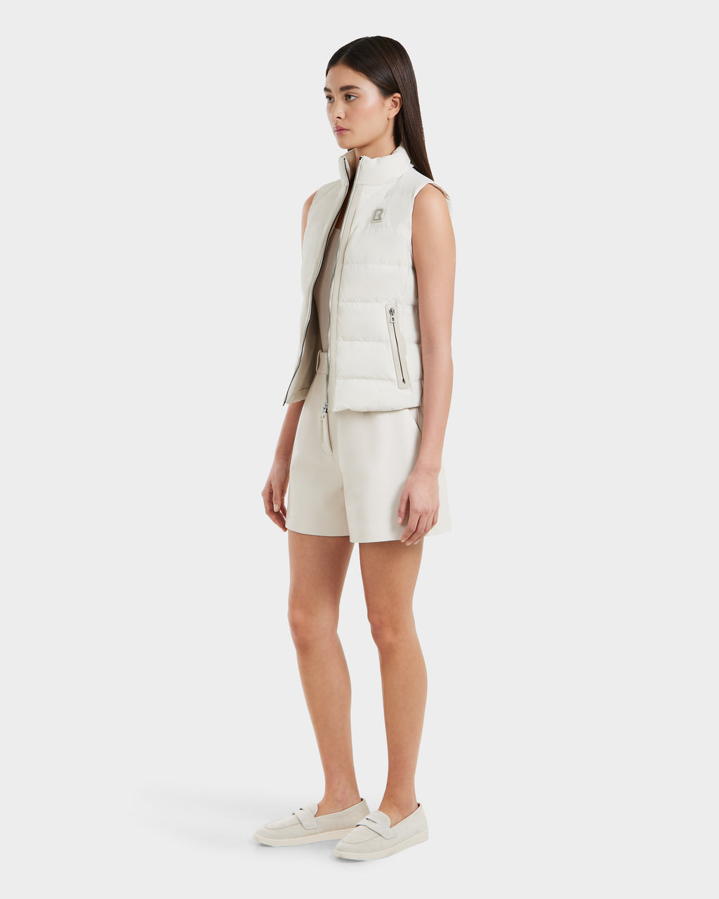 A woman wears the Rudsak GABRIELA - 8125014 Vanilla women's down puffer vest with vegan leather trim over a cream shirt and matching shorts, paired with white slip-on shoes, against a plain white background.