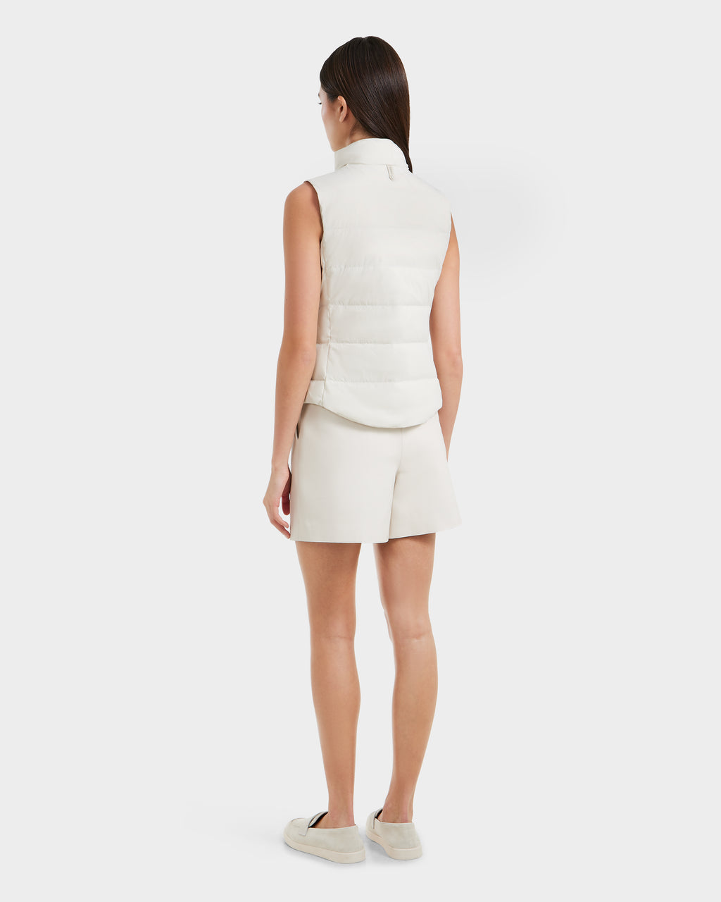 A woman with straight brown hair faces away, wearing the Rudsak GABRIELA - 8125014 Vanilla sleeveless down puffer vest with vegan leather trim, paired with white shorts and light slip-on shoes, against a plain white background.