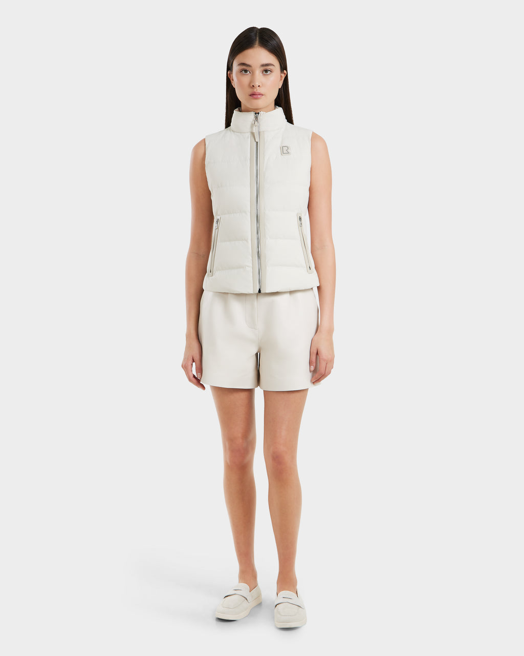 A woman faces forward wearing the Rudsak GABRIELA - 8125014 Vanilla, a white lightweight puffer vest with vegan leather trim, paired with white shorts and slip-on shoes against a plain, light background.