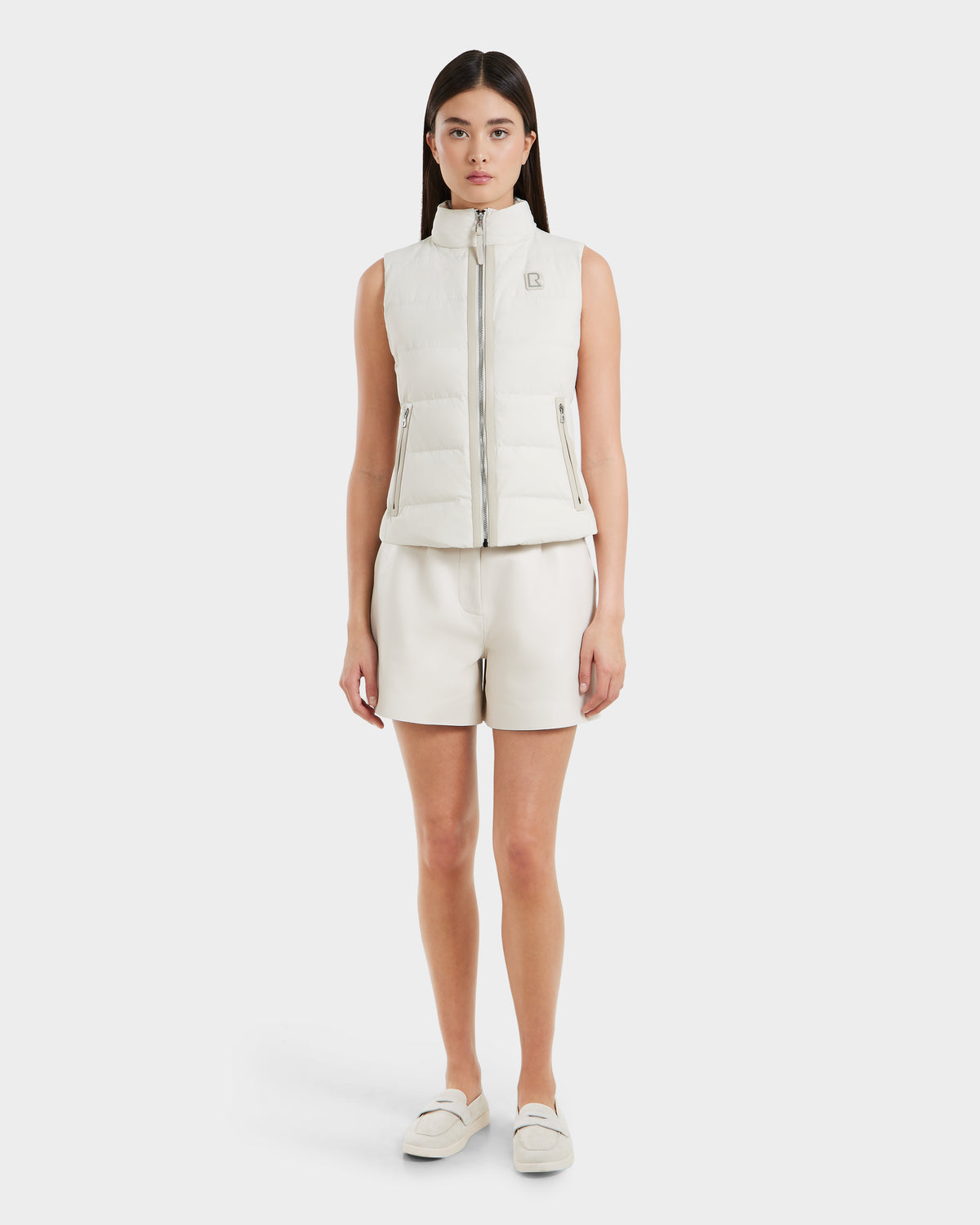 A woman faces forward wearing the Rudsak GABRIELA - 8125014 Vanilla, a white lightweight puffer vest with vegan leather trim, paired with white shorts and slip-on shoes against a plain, light background.