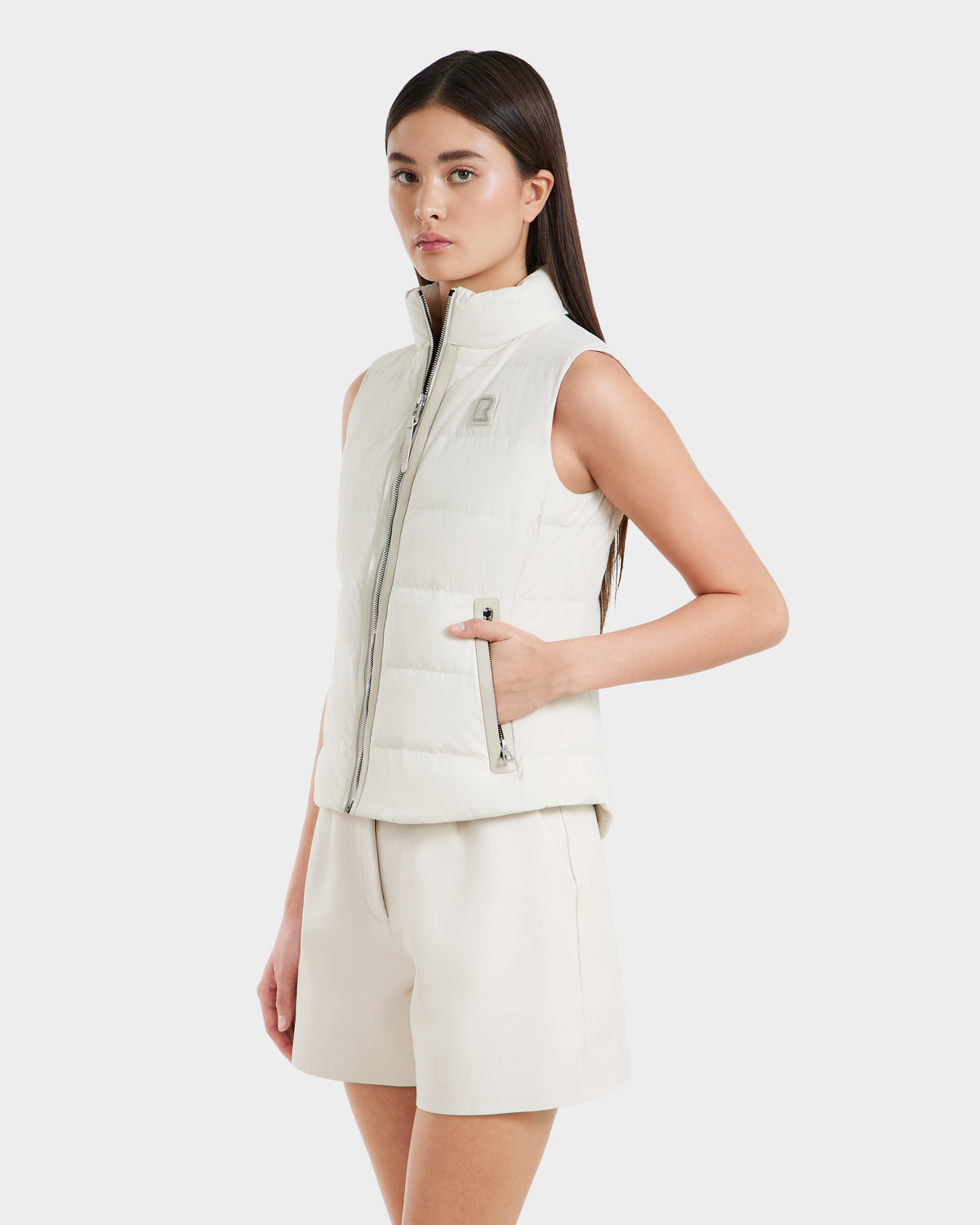 A woman with straight dark hair wears the Rudsak GABRIELA - 8125014 Vanilla sleeveless down puffer vest with vegan leather trim and light shorts, posing against a plain white background.