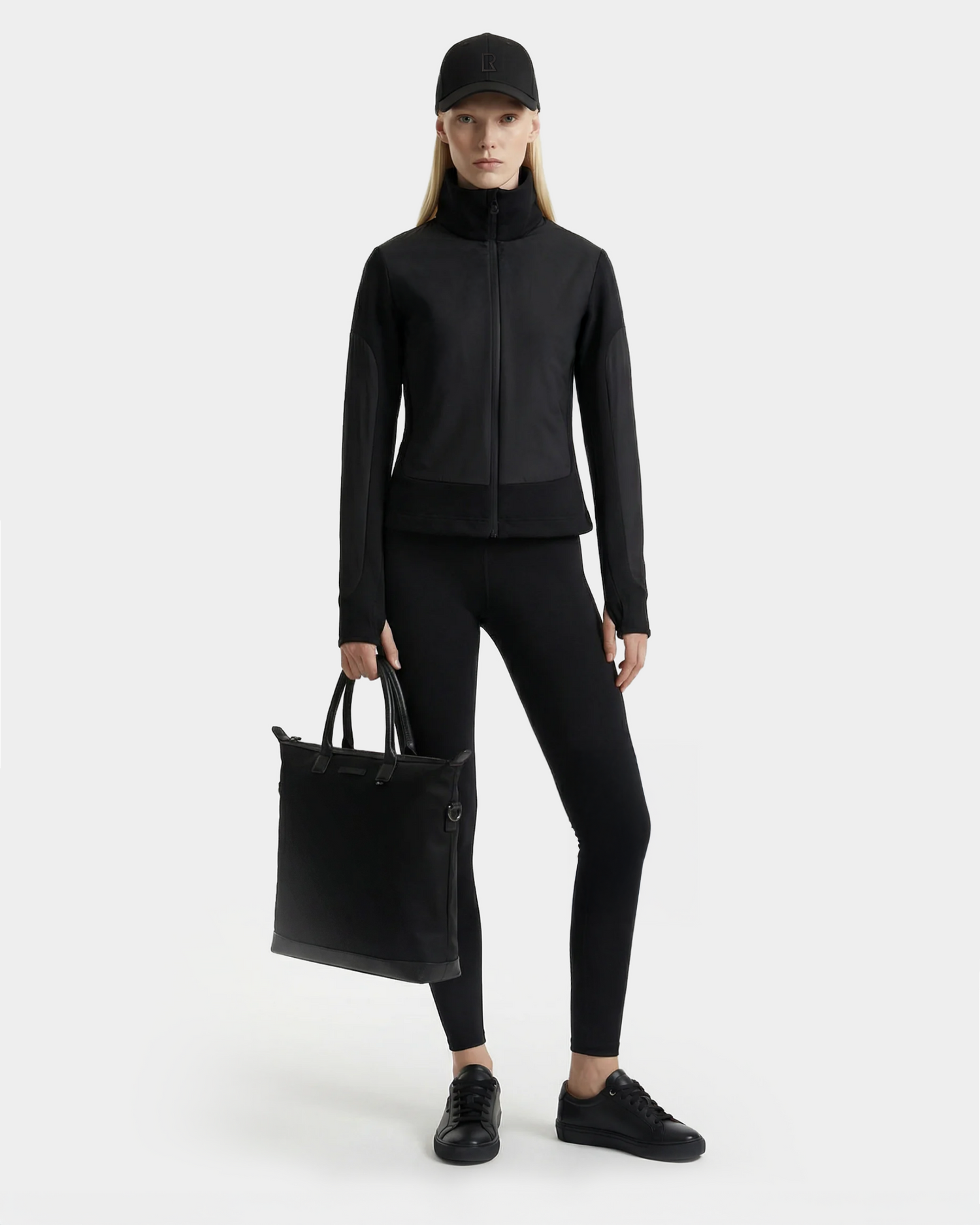 A woman faces forward in a sporty-chic all-black look, featuring the Rudsak GRANADA - 8126023 Black zip-up sweater jacket, leggings, sneakers, cap, and a black tote. The background is plain and light.