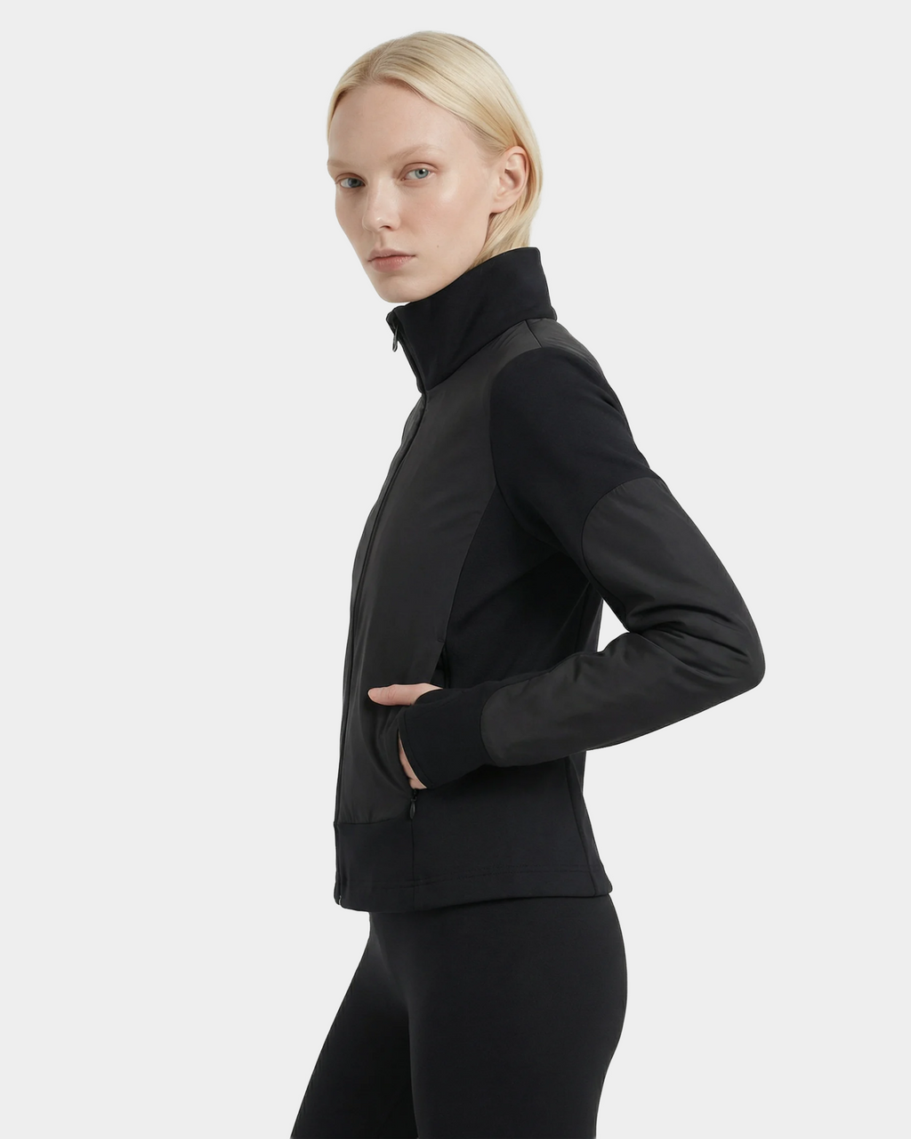 A person with blonde hair wears the Rudsak GRANADA - 8126023 Black fitted zip-up sweater and black pants, standing in profile with one hand in their jacket pocket against a plain light background.