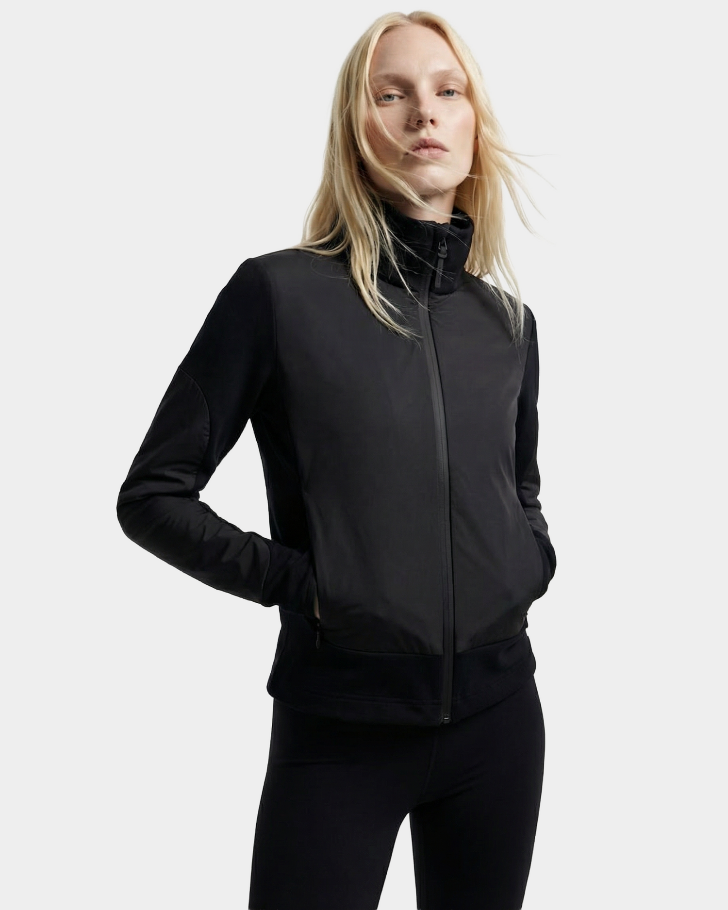 A person with long blonde hair models the Rudsak GRANADA - 8126023 Black fitted lightweight jacket and black pants, standing hands-in-pockets against a plain light gray background.