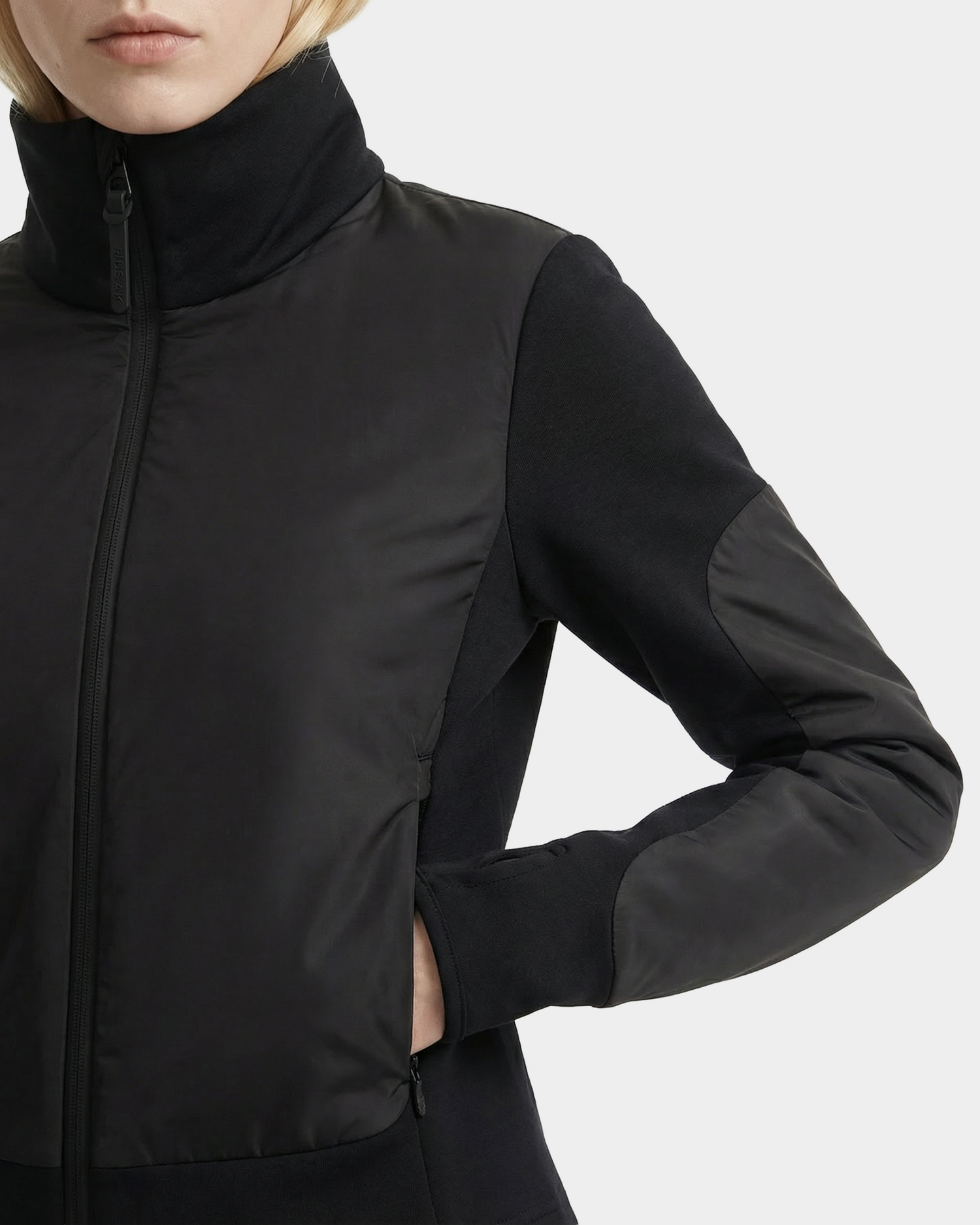 A woman wears the Rudsak GRANADA - 8126023 Black high-collar zip-up sweater jacket with her left hand in the pocket, shown from the chest up against a plain background.