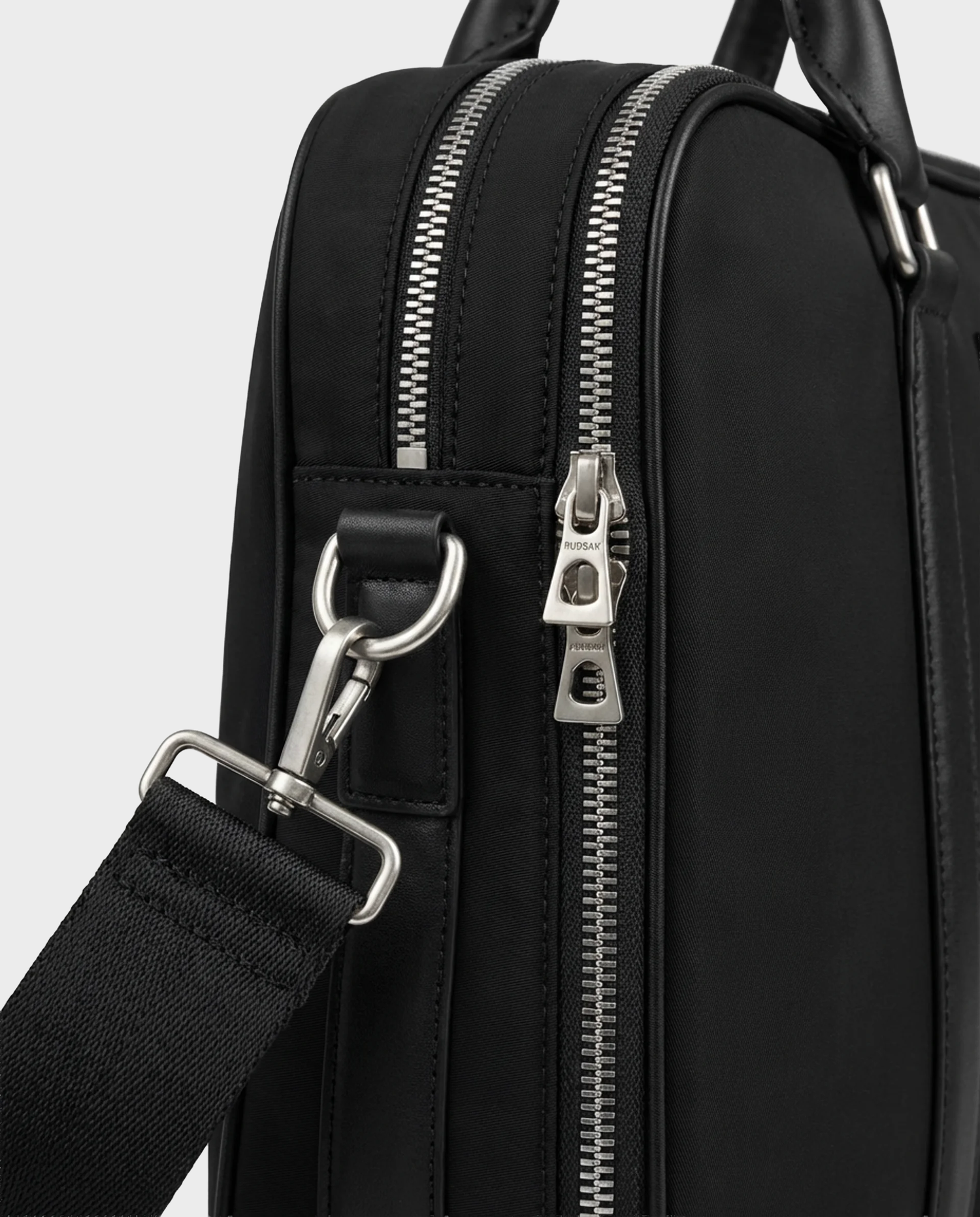 Close-up of the Rudsak HENRY N - 9326056 Black bag, featuring silver zippers, a shoulder strap attached with a metal clasp, and detailed stitching that highlights the textured material and hardware.