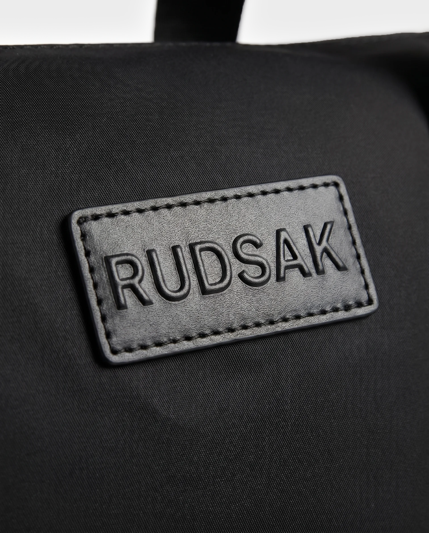A close-up of a black fabric patch embossed with “Rudsak”, stitched onto the surface of the HENRY N - 9326056 Black by Rudsak.