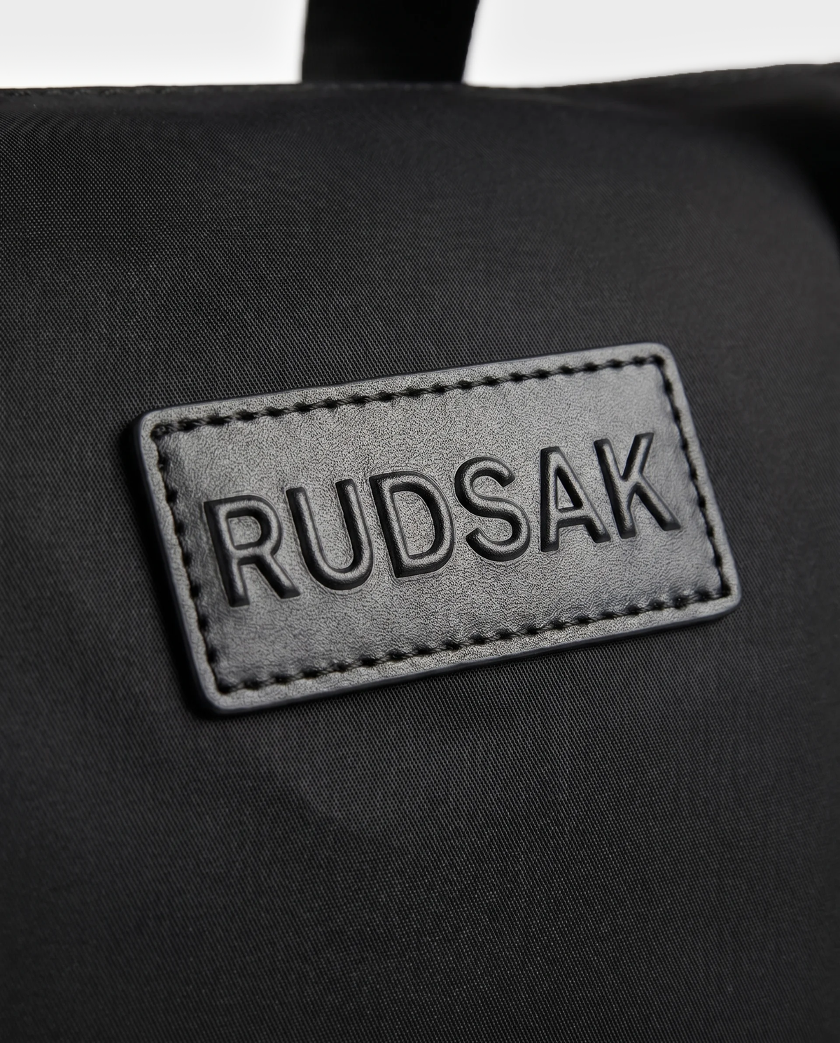 A close-up of a black fabric patch embossed with “Rudsak”, stitched onto the surface of the HENRY N - 9326056 Black by Rudsak.