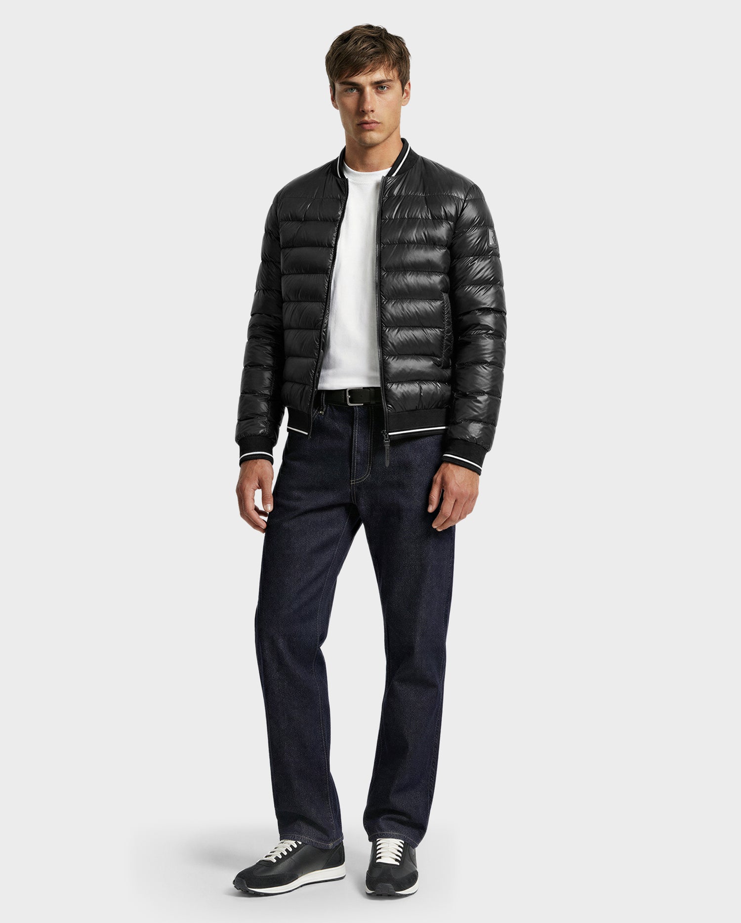 A man faces forward wearing the Rudsak JEREMY - 6125025 Black men’s down bomber jacket, a white T-shirt, dark blue jeans, and gray sneakers against a plain light gray background.